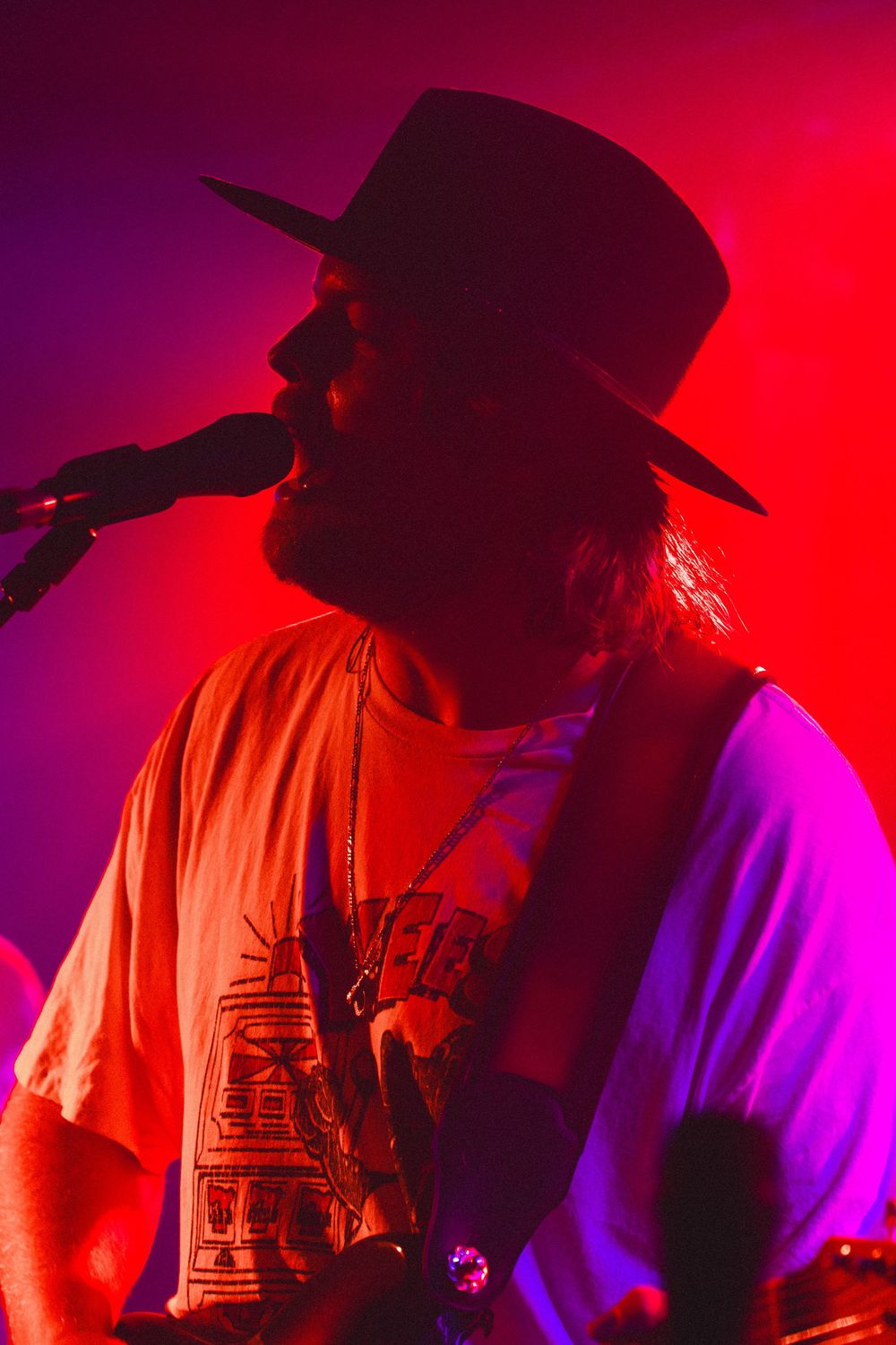 Silhouetted male singer wearing a wide-brim hat performs at a live concert under dramatic red and pink stage lighting.