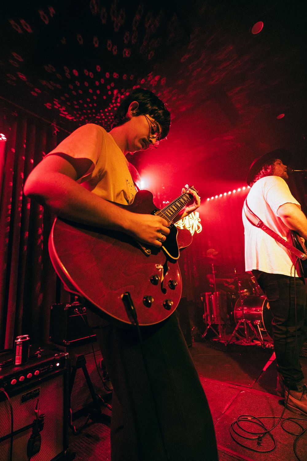 Young guitarist wearing glasses plays a red semi-hollow electric guitar on a red-lit concert stage.