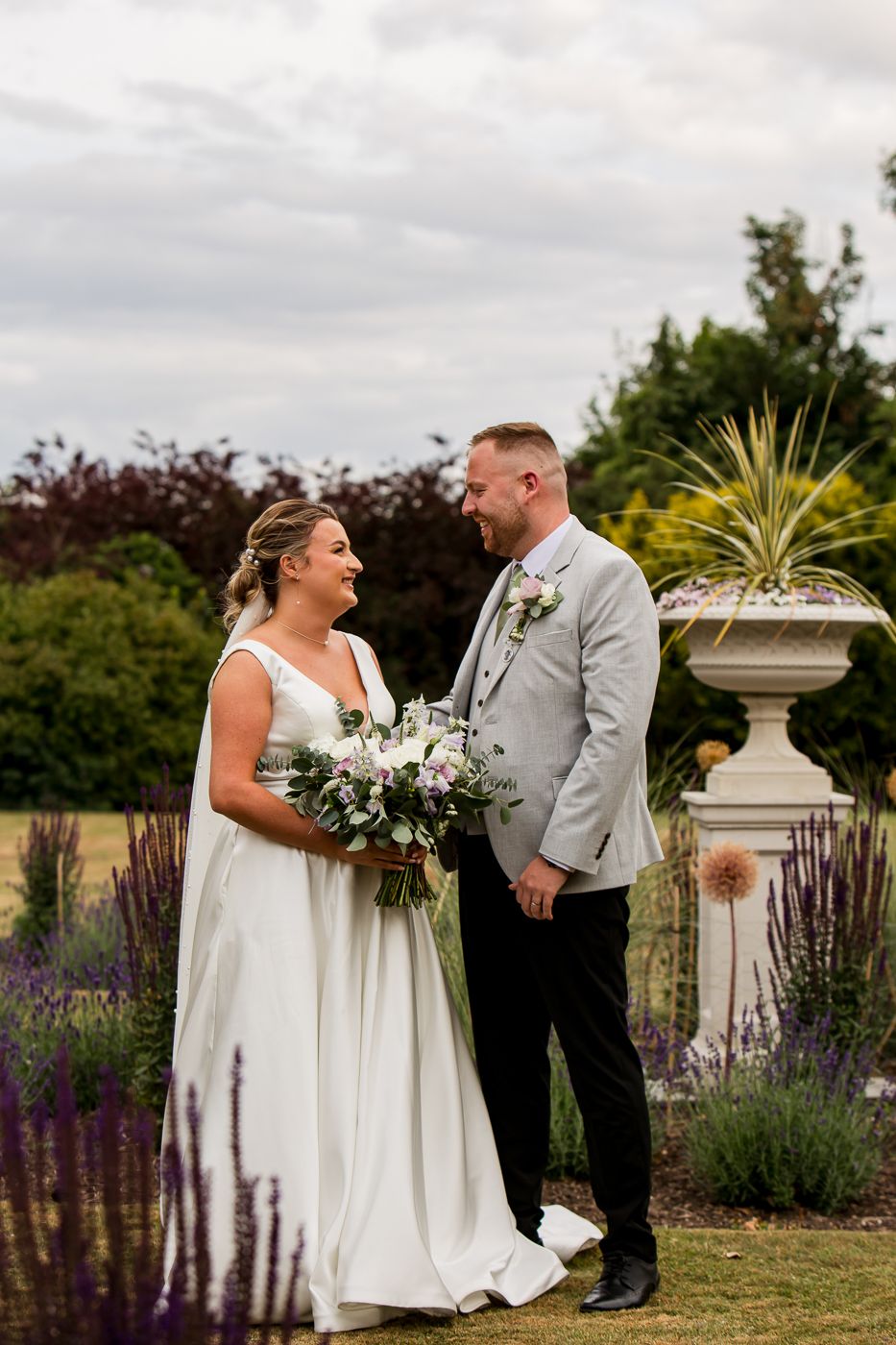 A wedding couple poses together in a garden with purple flowers and a decorative stone fountain.