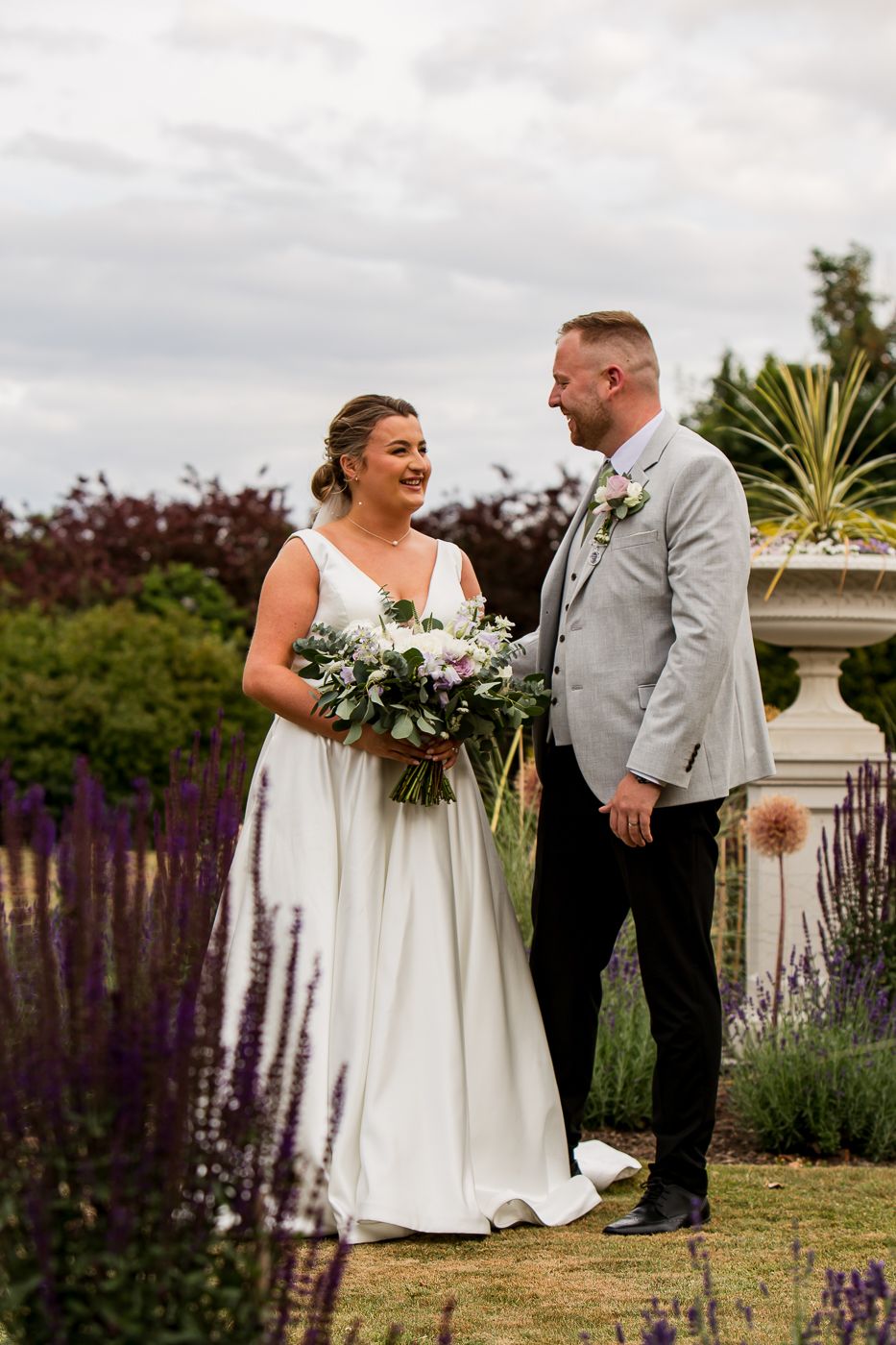 A romantic garden wedding scene with the couple standing among purple flowers and greenery.