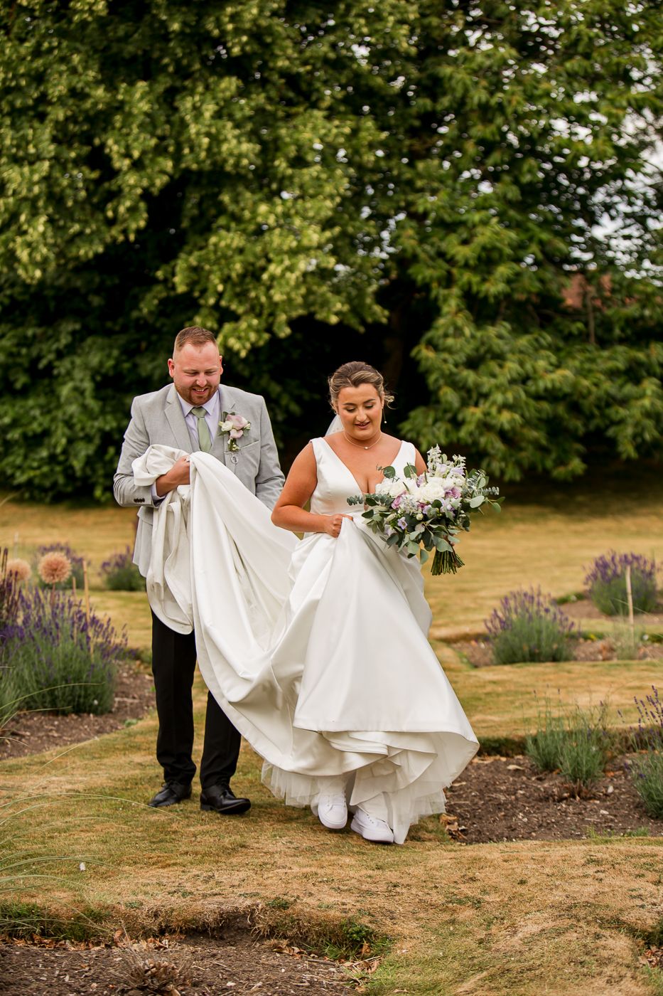 A couple walks through a garden path with the bride lifting her white dress.