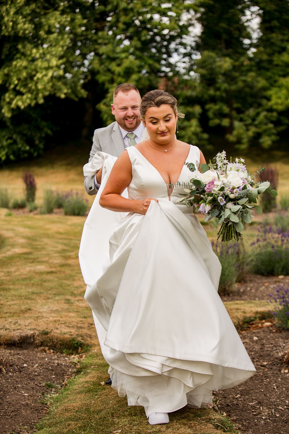 A bride in a flowing white dress lifts her skirt while walking through a garden with her partner.