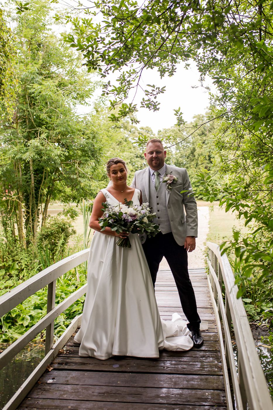 A couple poses on a wooden bridge surrounded by lush green trees during their wedding photoshoot.