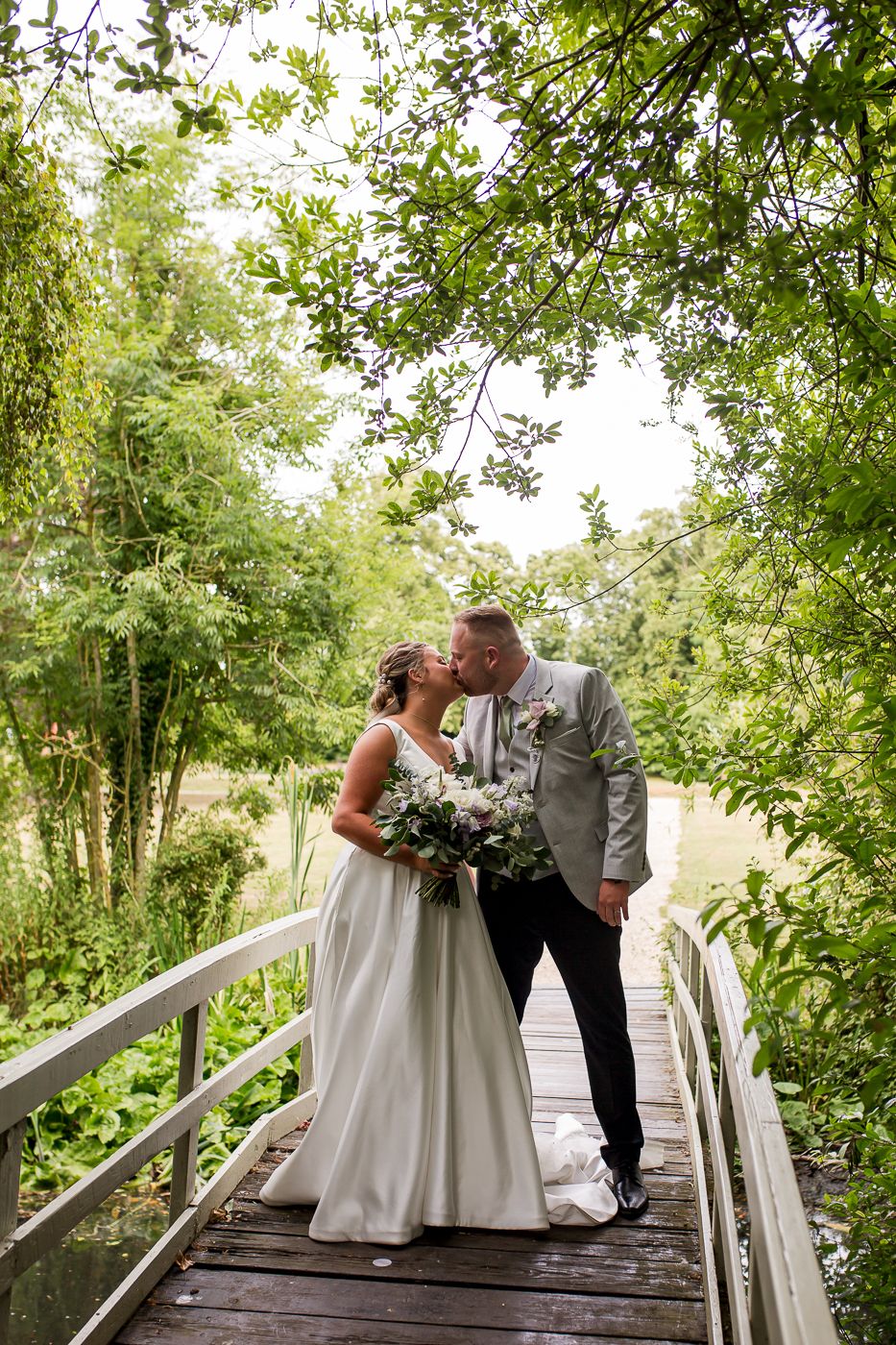 A wedding couple stands together on a wooden bridge pathway surrounded by trees.