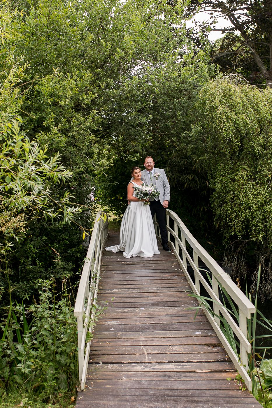 A wooden footbridge surrounded by lush green foliage creates a romantic setting for wedding photos.