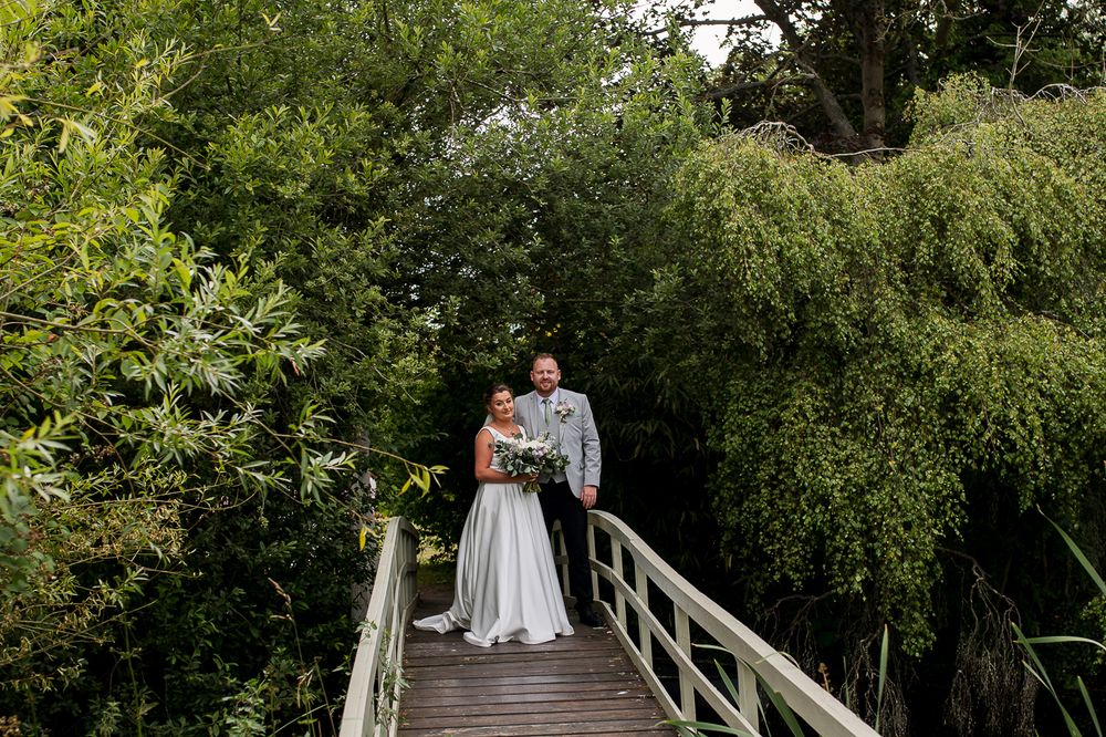 Wedding couple stands on a rustic wooden bridge surrounded by verdant summer greenery.