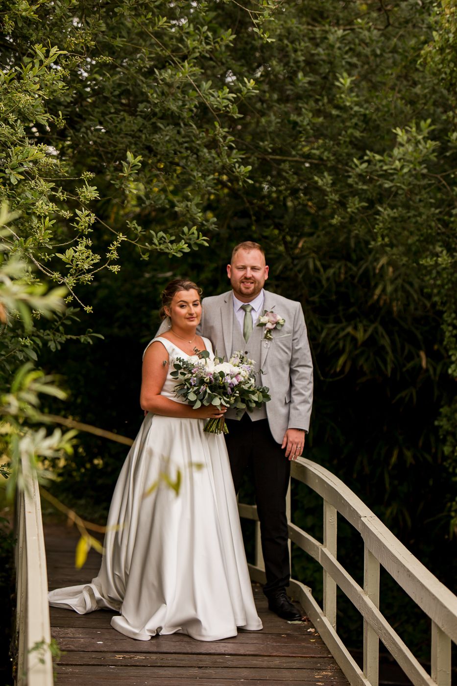 Newlyweds share an intimate moment on a wooden bridge deck amidst leafy trees.