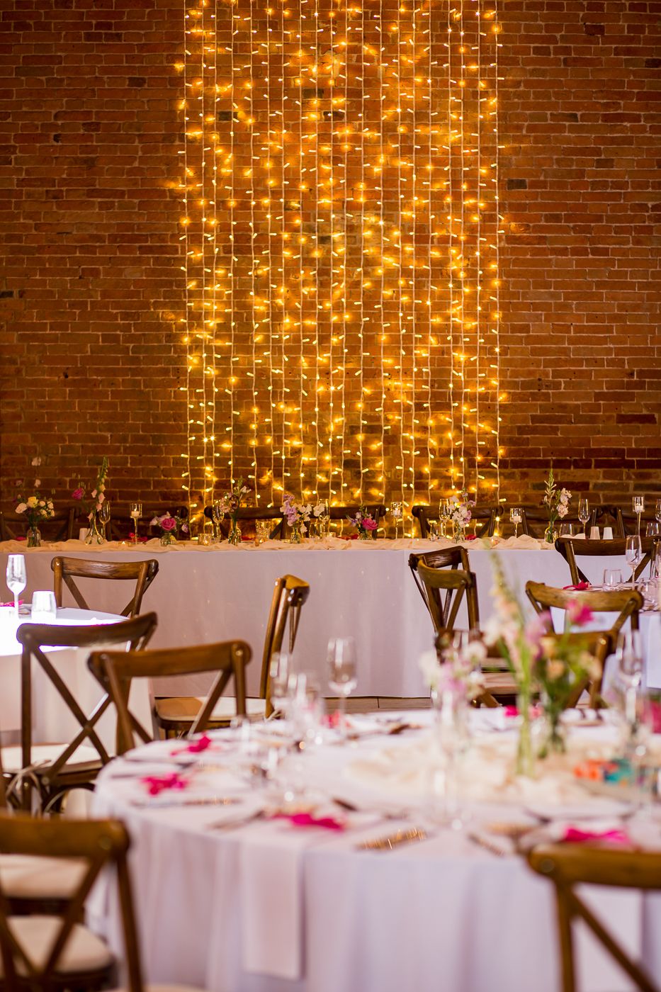 Elegant wedding reception tables with white linens and wooden chairs against a brick wall with cascading fairy lights.
