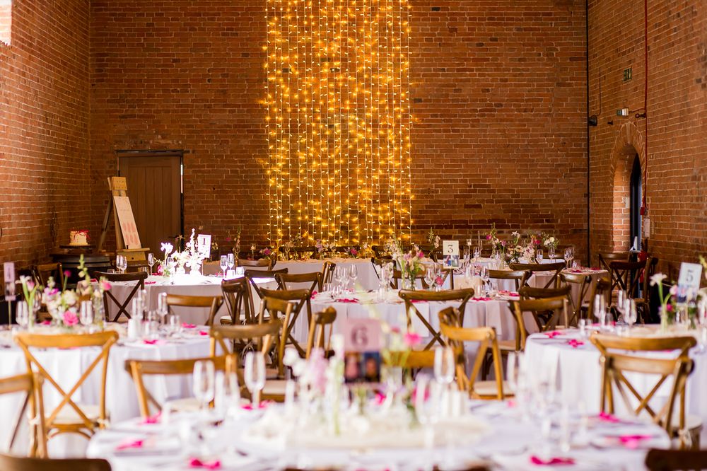Wedding venue with round tables and crossback chairs illuminated by a dramatic wall of string lights.