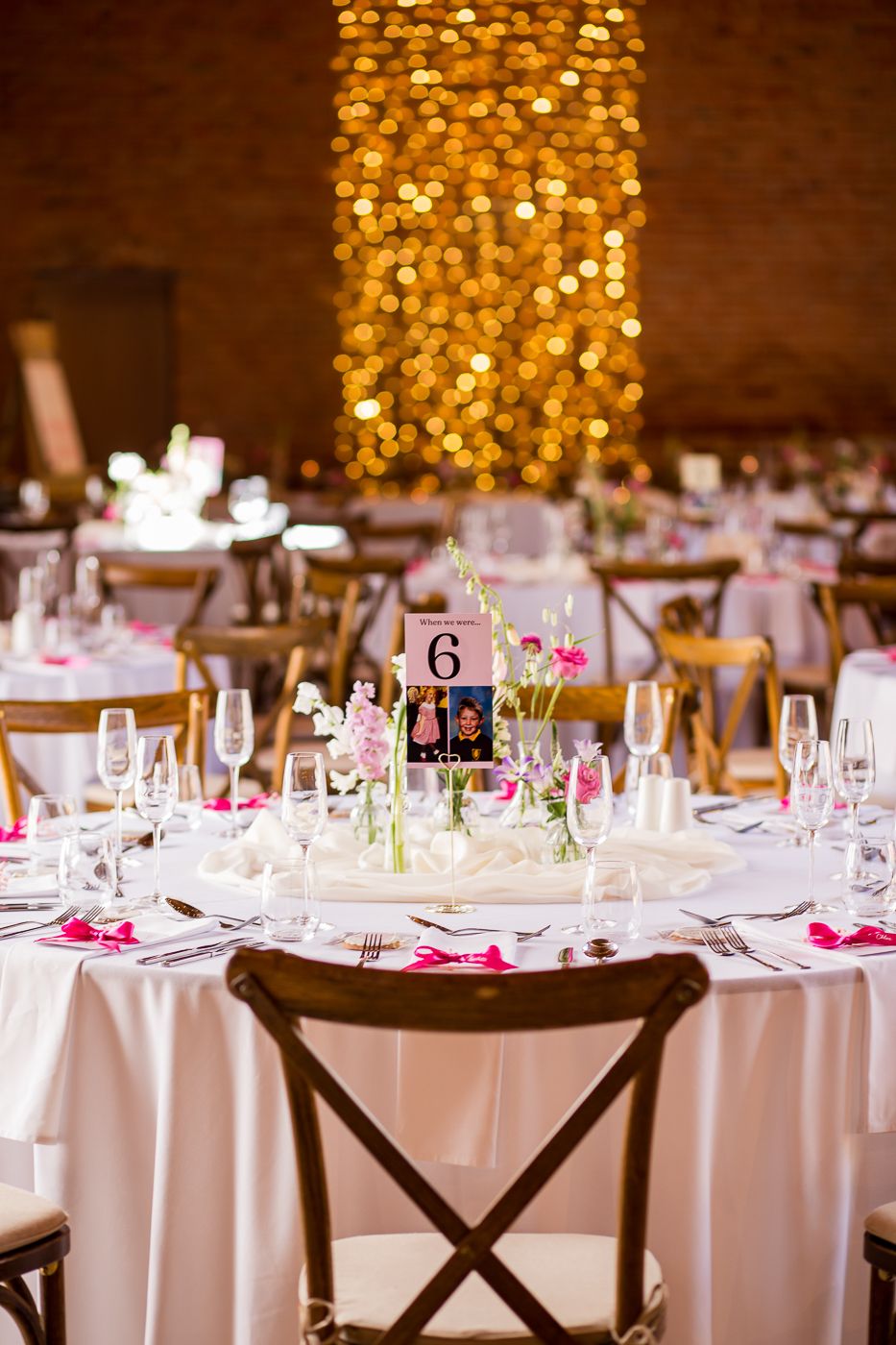 Round wedding reception table with white linens and pink floral centerpieces under golden fairy lights.