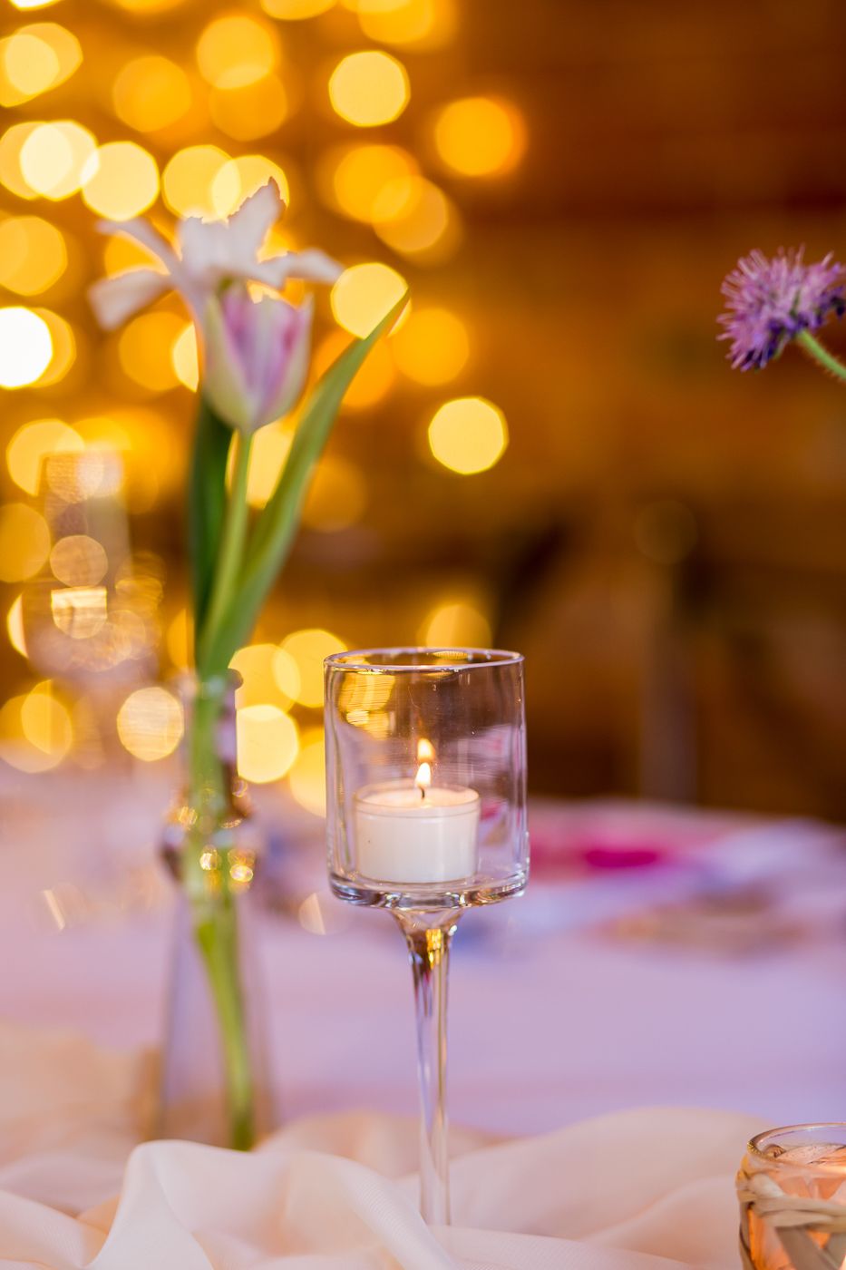 Romantic table setting with iris flower in vase and stemmed candle holder against bokeh lights.