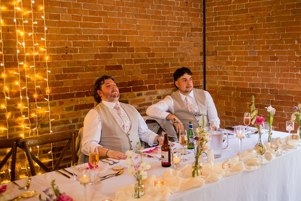 Wedding reception head table decorated with string lights and white linens against brick wall.