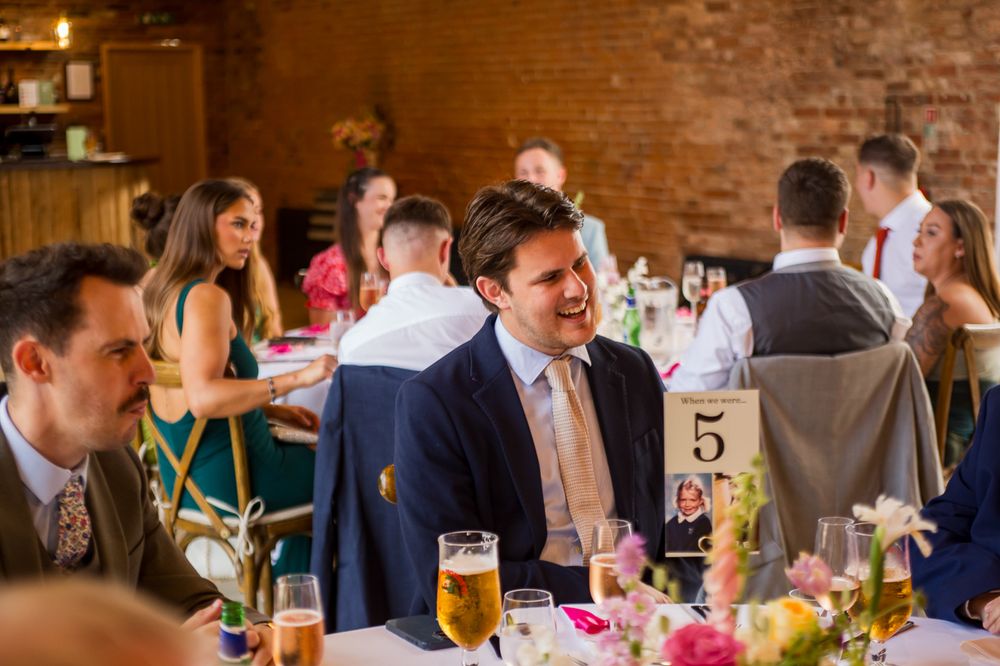 Wedding guests seated at long tables in a rustic brick venue with string lights overhead during dinner reception.