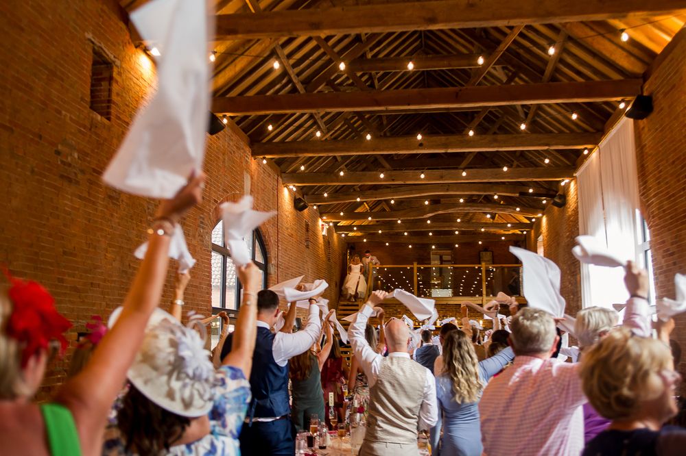 Wedding guests waving napkins in celebration inside a rustic brick venue with string lights.