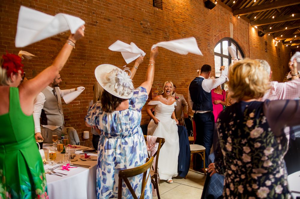 Celebratory napkin waving during wedding reception in rustic brick venue with string lights.
