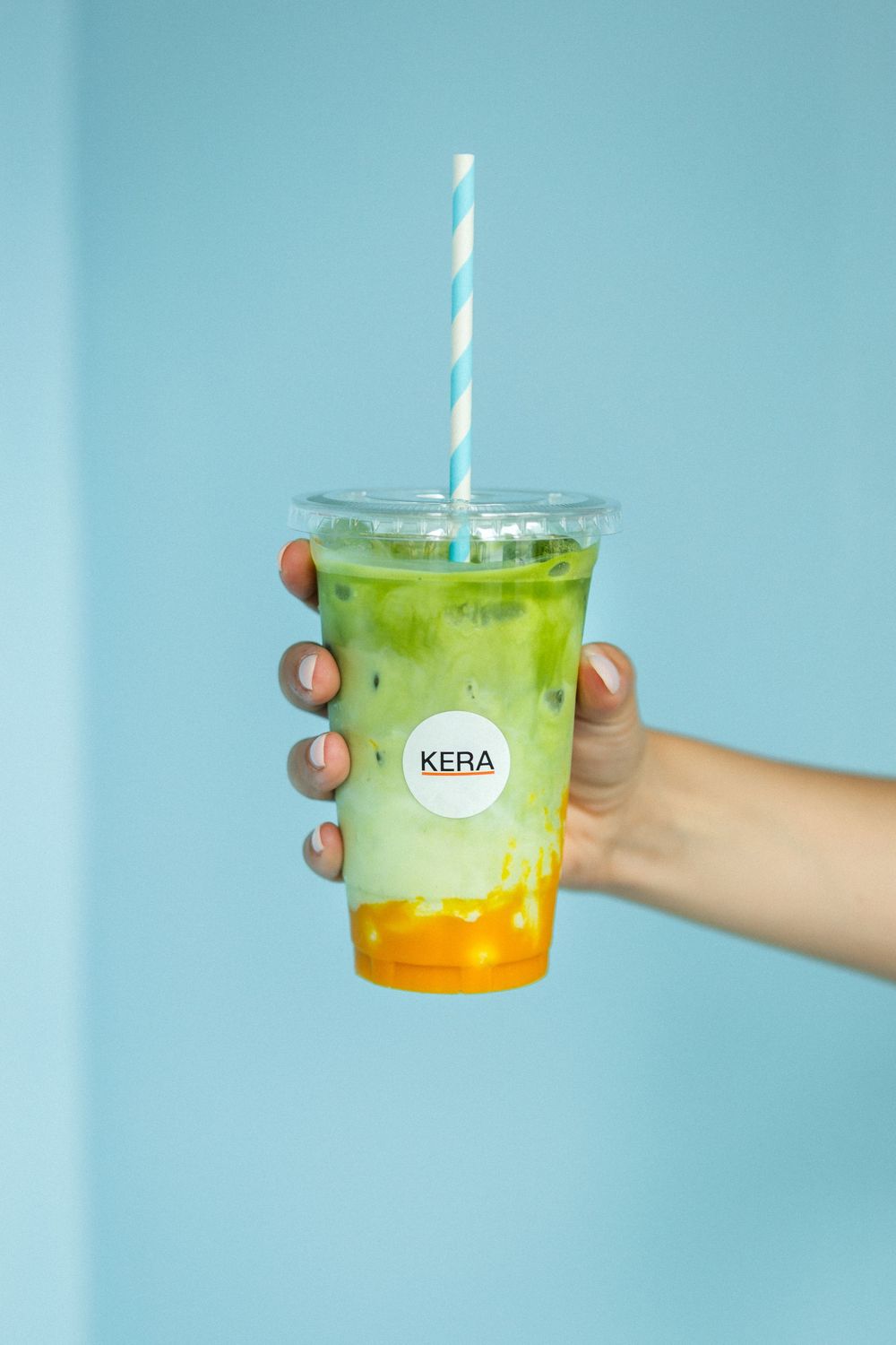 A hand holds a plastic cup of layered green matcha drink with orange syrup and a striped straw against a blue background.