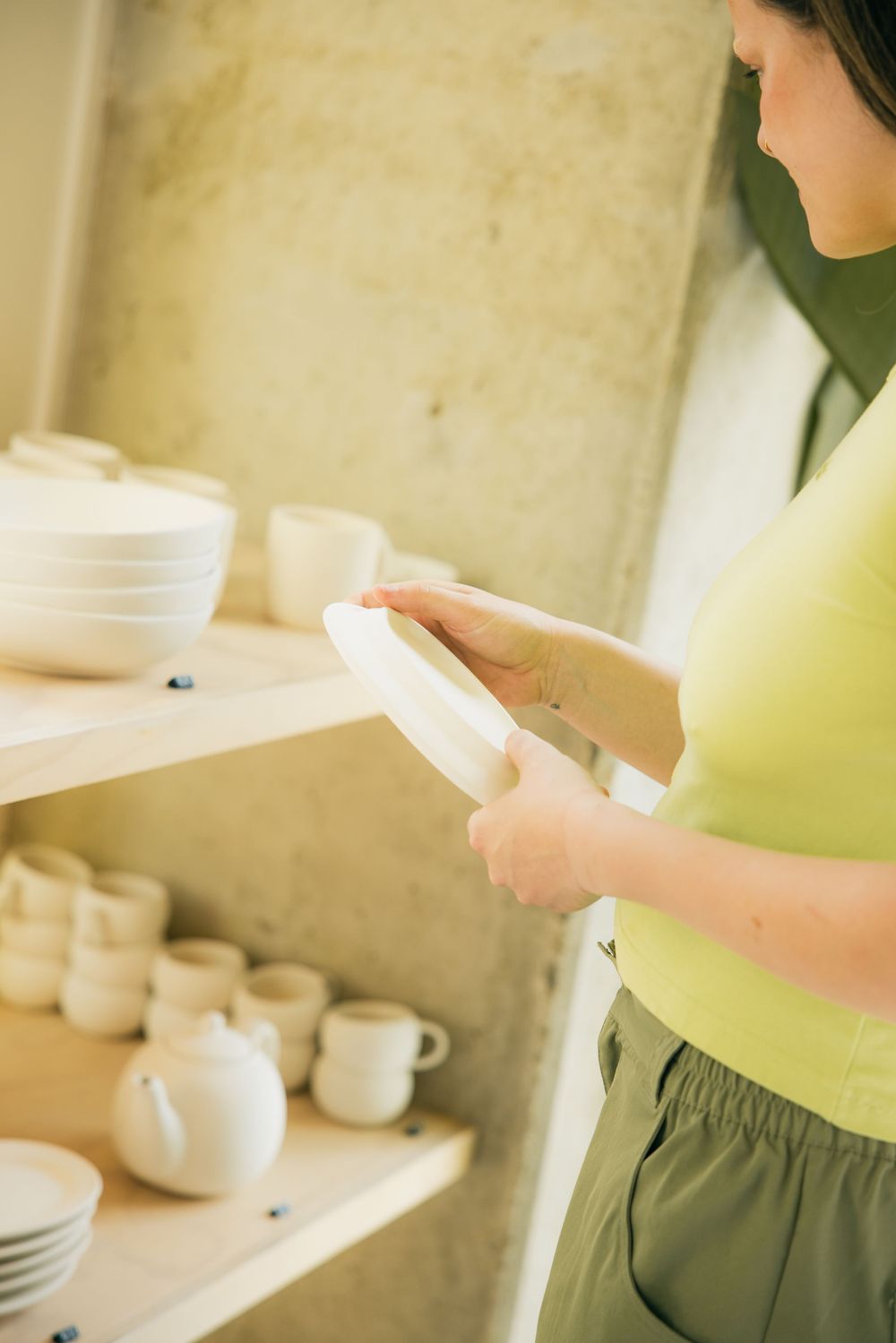 Woman in yellow top examining white ceramic plate near shelves of pottery cups and bowls in a bright shop.