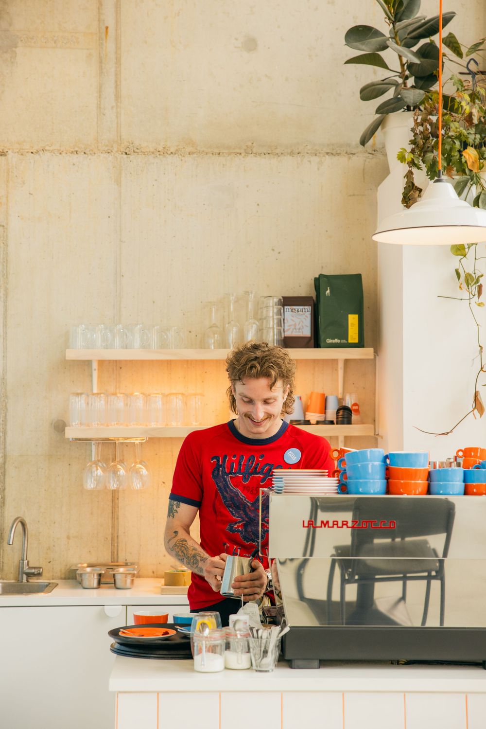 Tattooed barista in red t-shirt smiling while operating an espresso machine in a modern cafe.