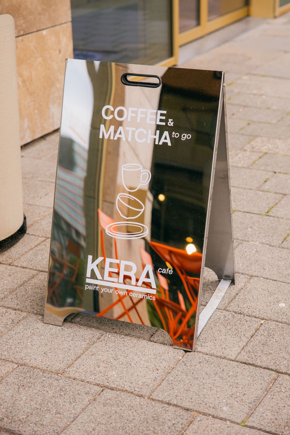 Shiny metallic A-frame sign for Kera Cafe advertising coffee and matcha to go on a paved sidewalk.