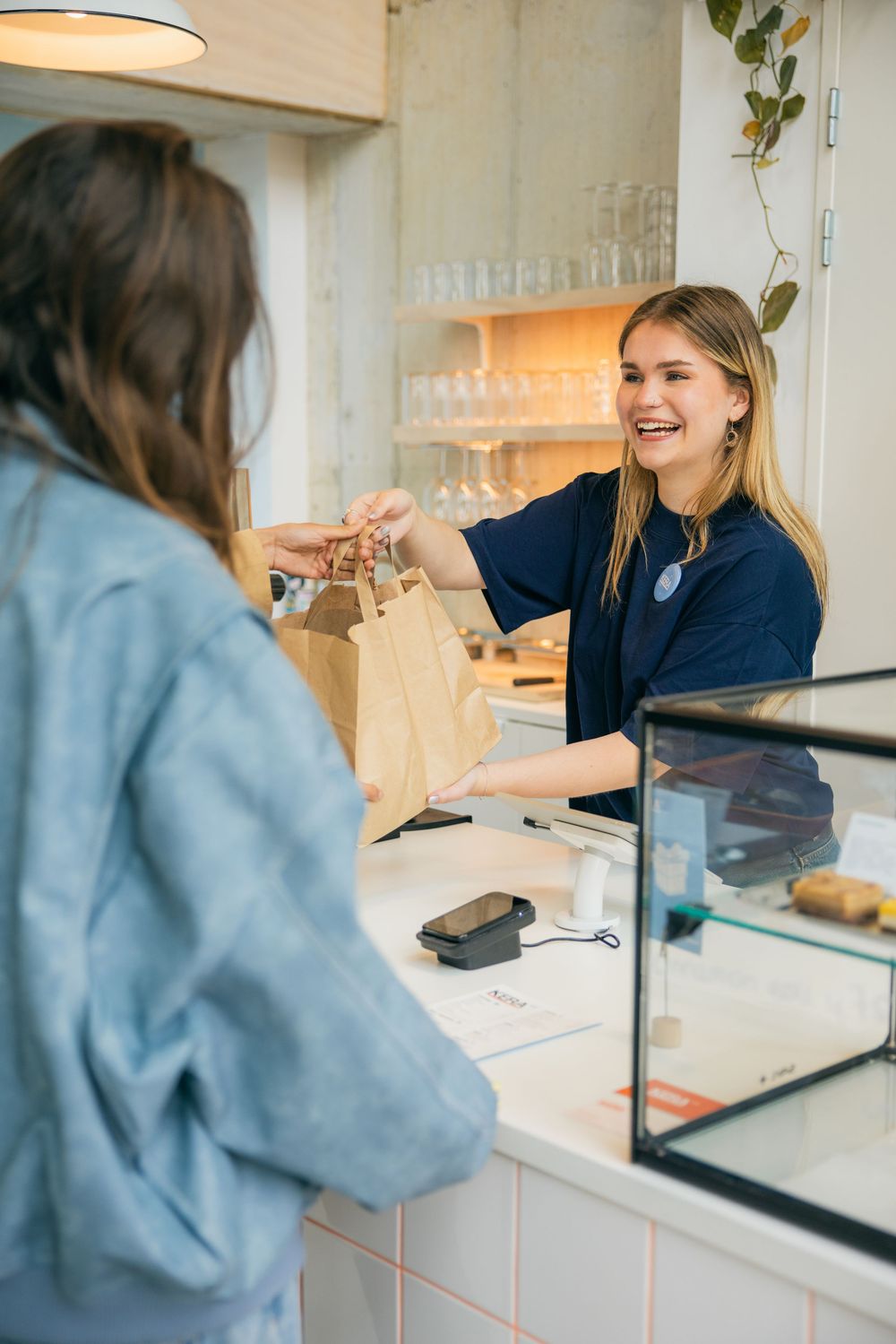 Smiling female barista handing a paper bag to a customer at a modern cafe counter.