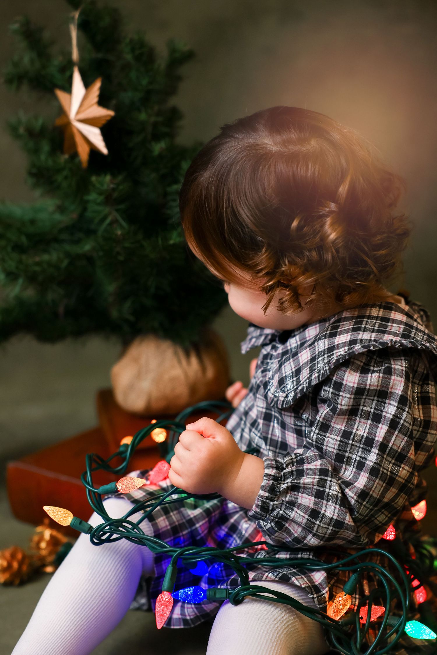 A small child in plaid lies on a white surface with colorful Christmas lights creating a festive holiday scene.