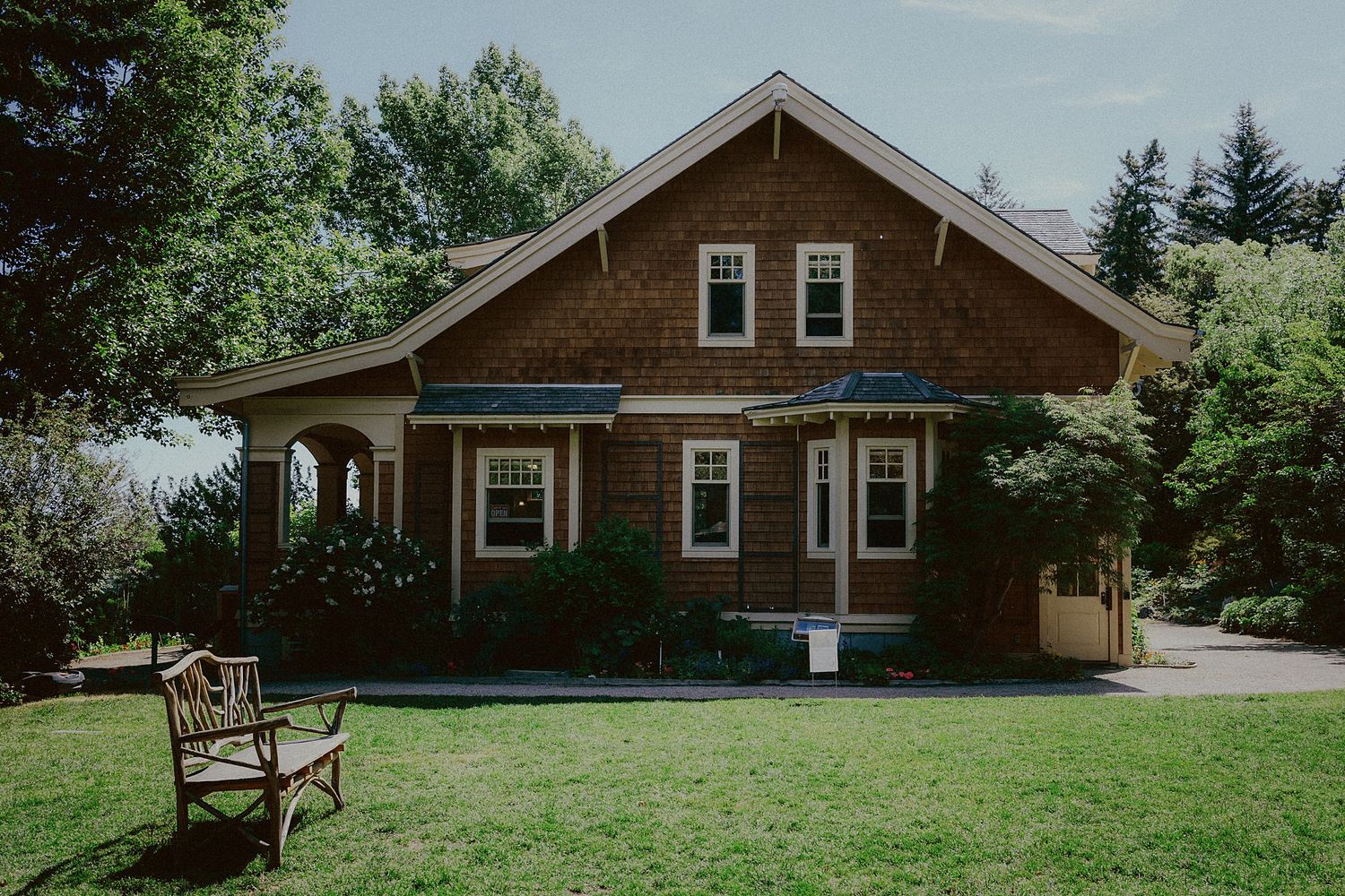 A charming brown craftsman style house with white trim and a covered porch sits on a green lawn with a wooden bench.