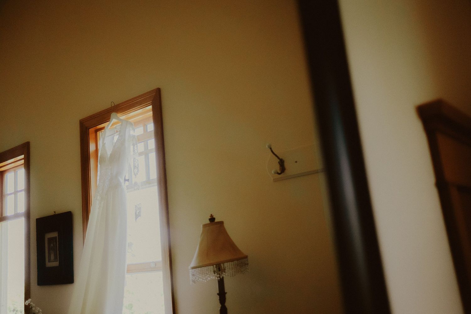 A white wedding dress hangs in a sunlit room with a floor lamp and curtained windows.