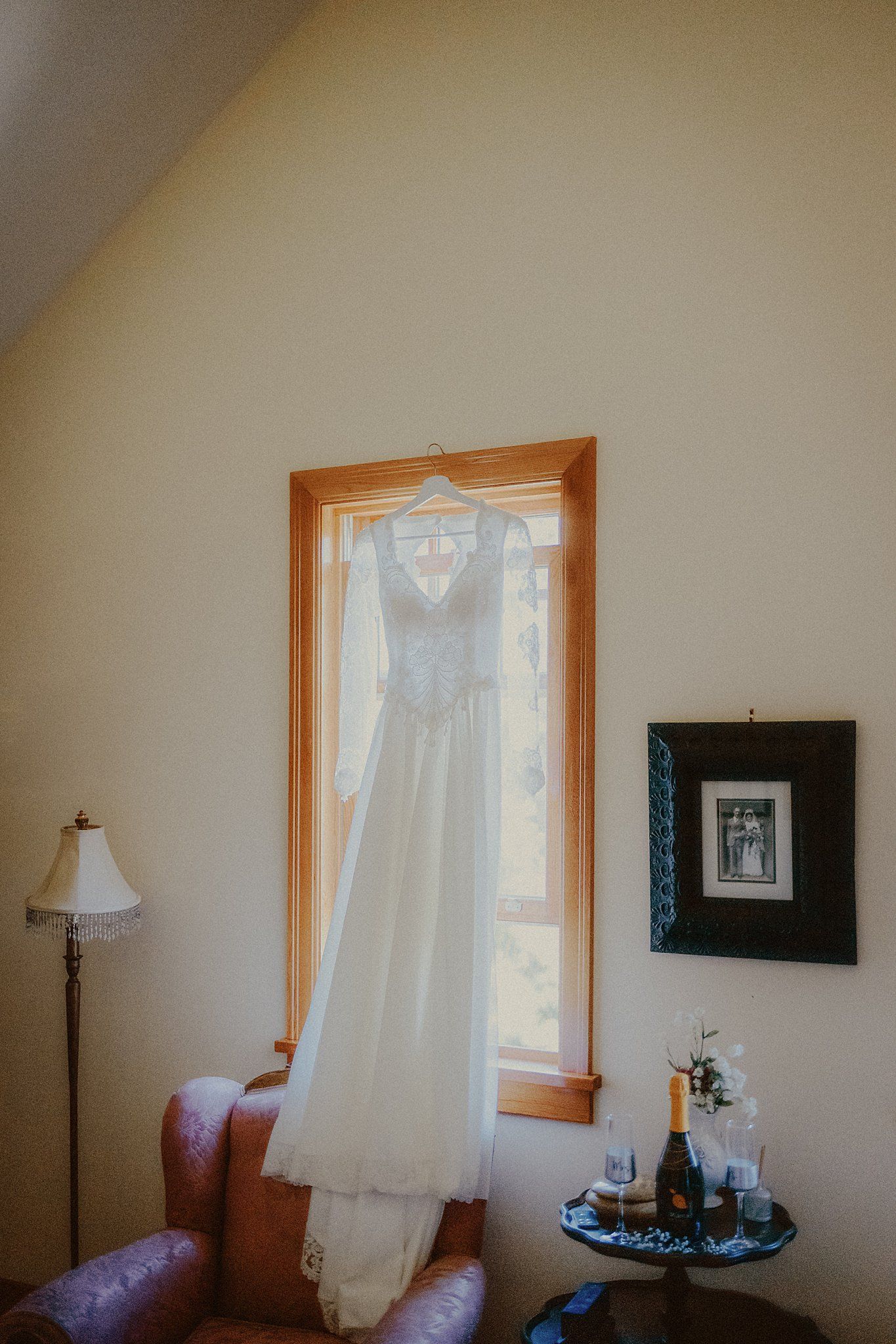A white wedding dress hangs in front of a window next to a floor lamp and small side table with decor.