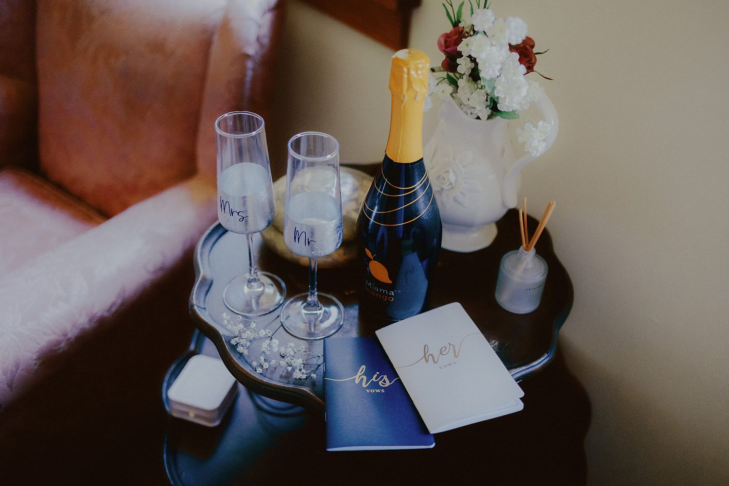 Wedding details including champagne, glasses, vases with flowers, and blue stationery arranged on a dark surface.