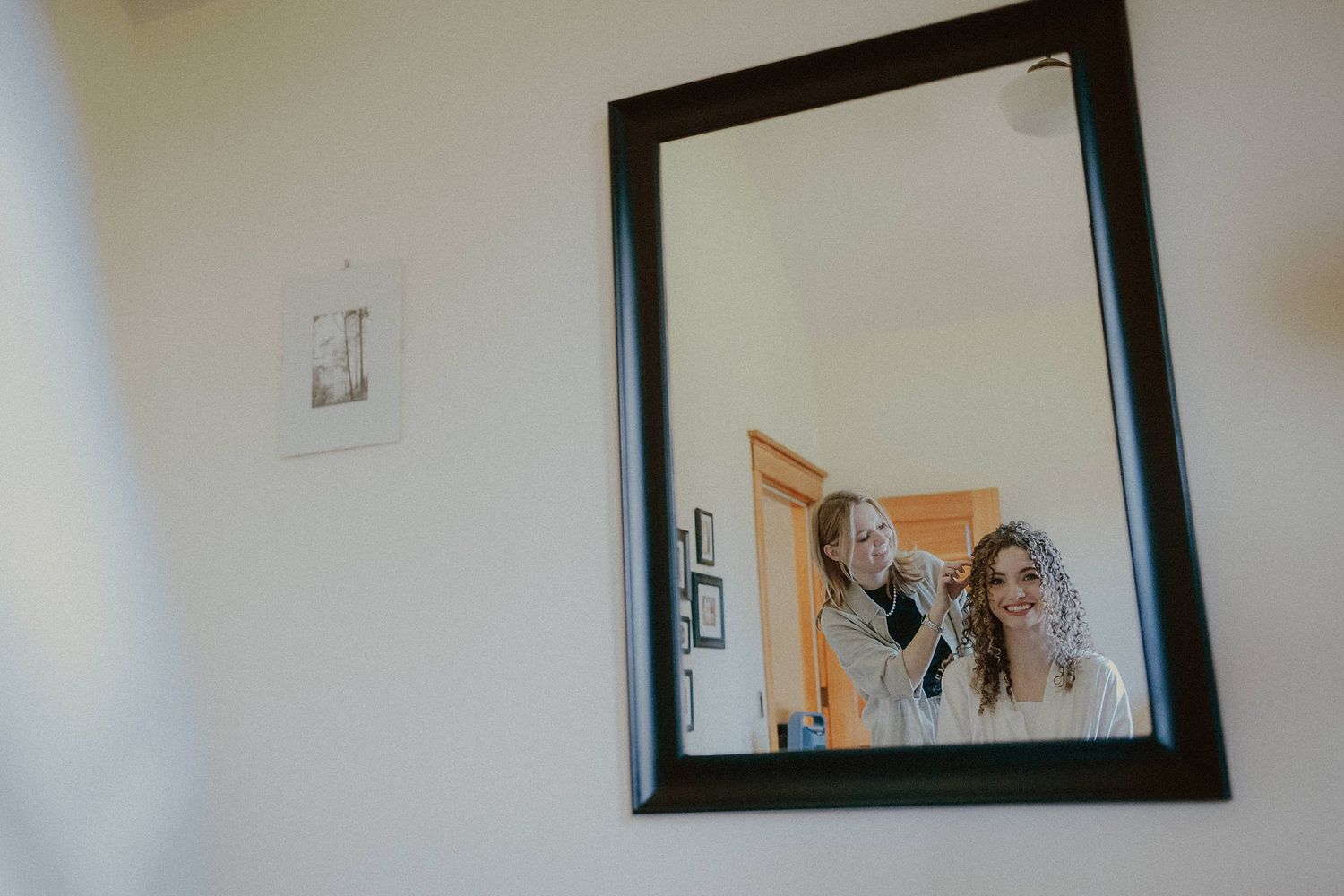 A reflection in a black-framed mirror mounted on a white wall shows wedding preparation moments.