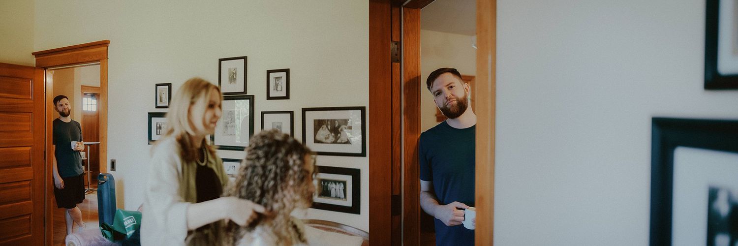 Two people stand in a hallway with framed photos on the wall, one looking in a mirror while the other observes.