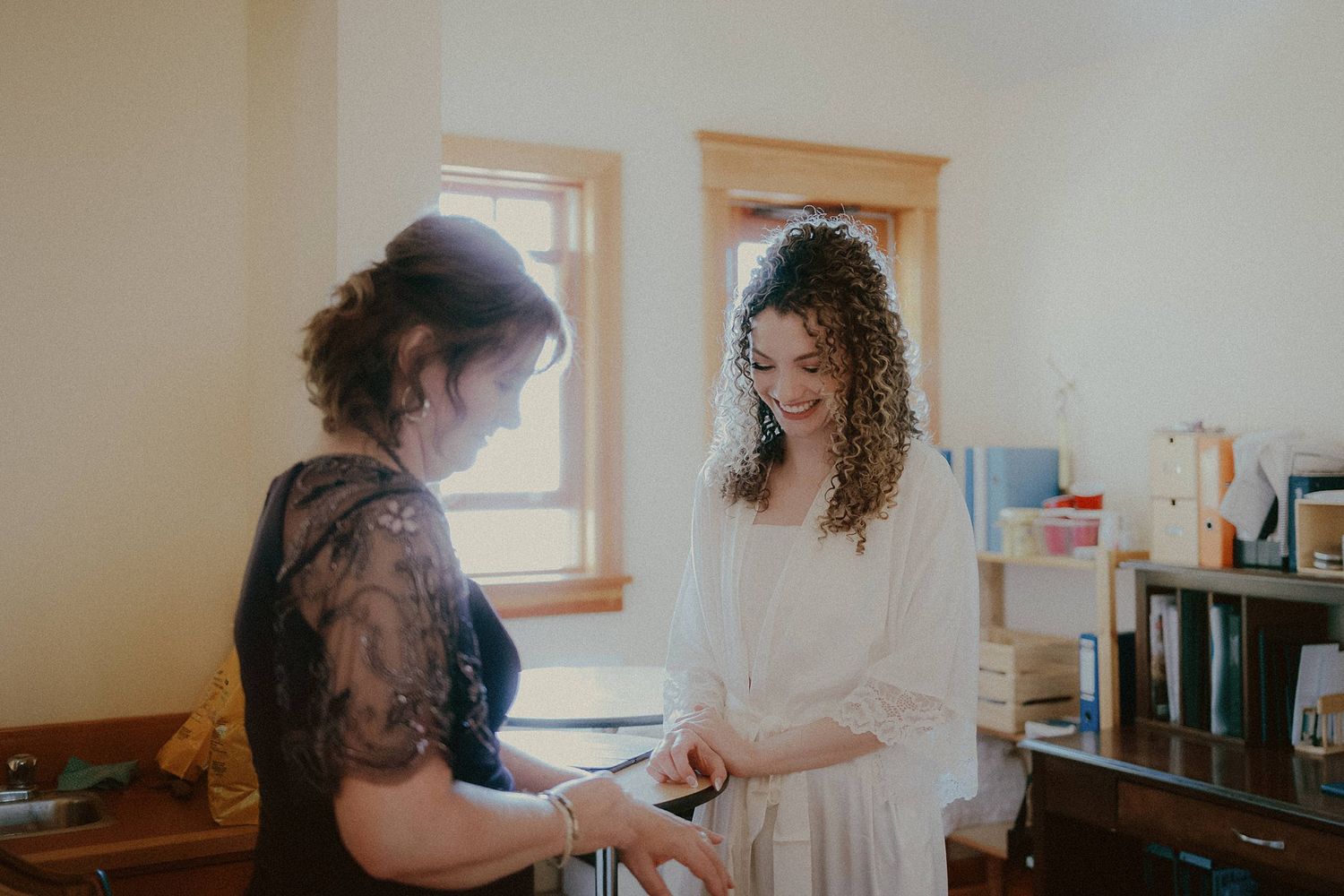 Two people share a tender moment during wedding preparations in a naturally lit room.