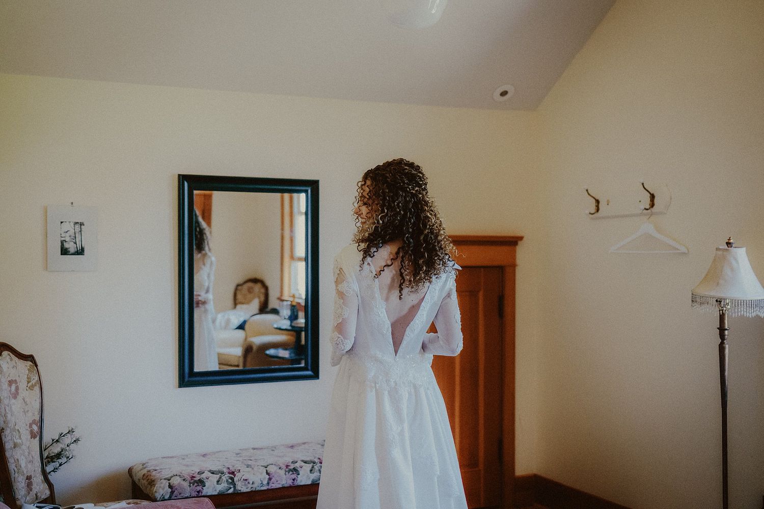 A wedding dress hangs in a sunlit room with framed photos on the wall and wooden furniture.