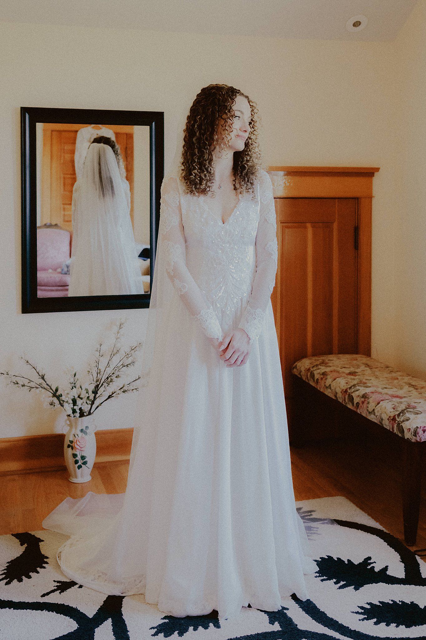 A white wedding dress with lace details hangs elegantly in a sunlit room with wooden furniture.