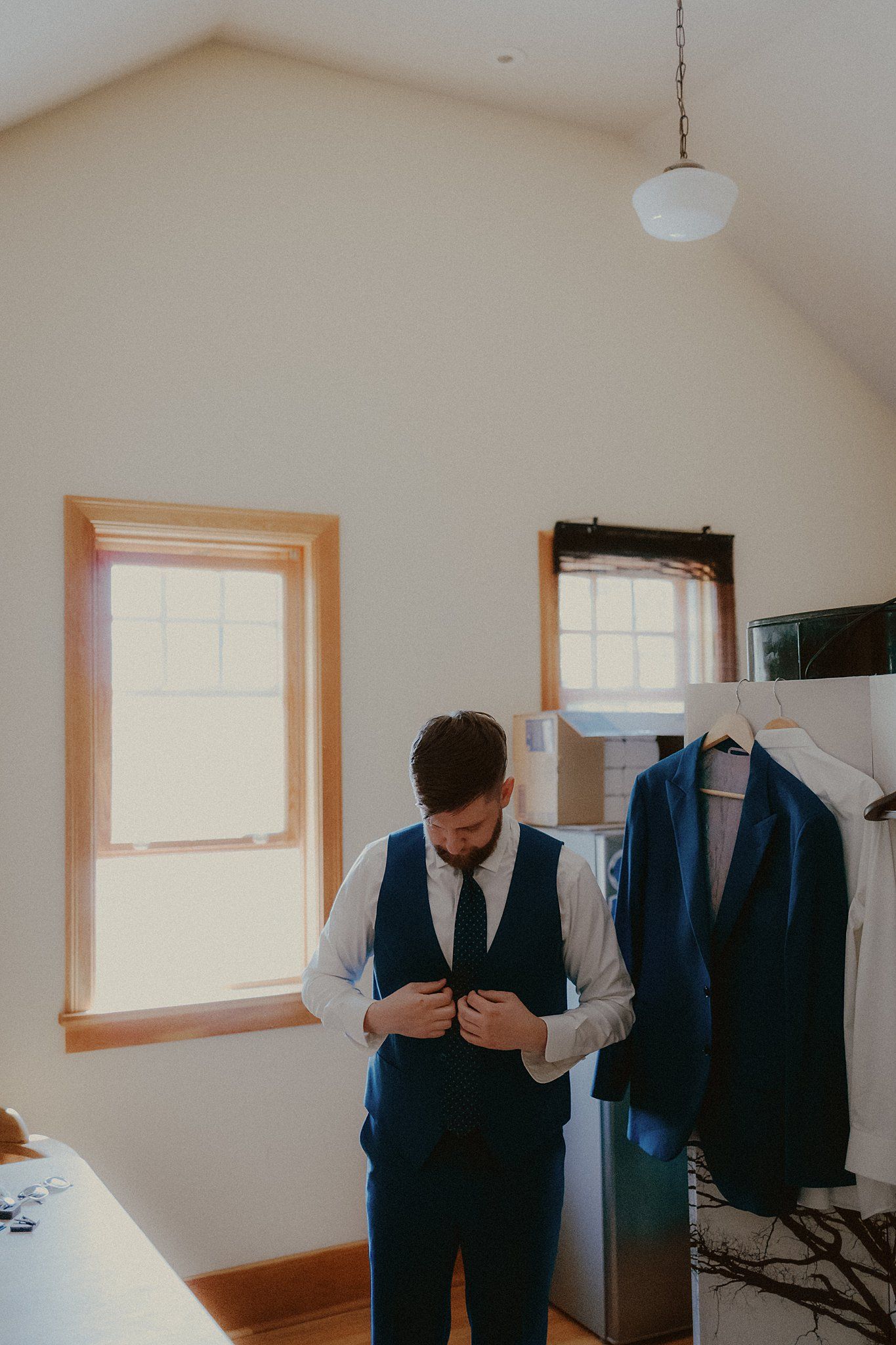 A groom adjusts his vest in a sunlit room with hanging formal attire.