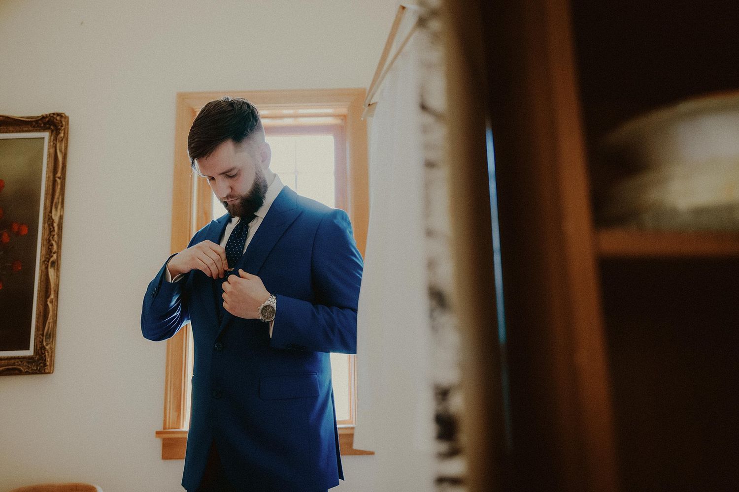 A person in a navy blue suit adjusts their tie while getting ready in a sunlit room.