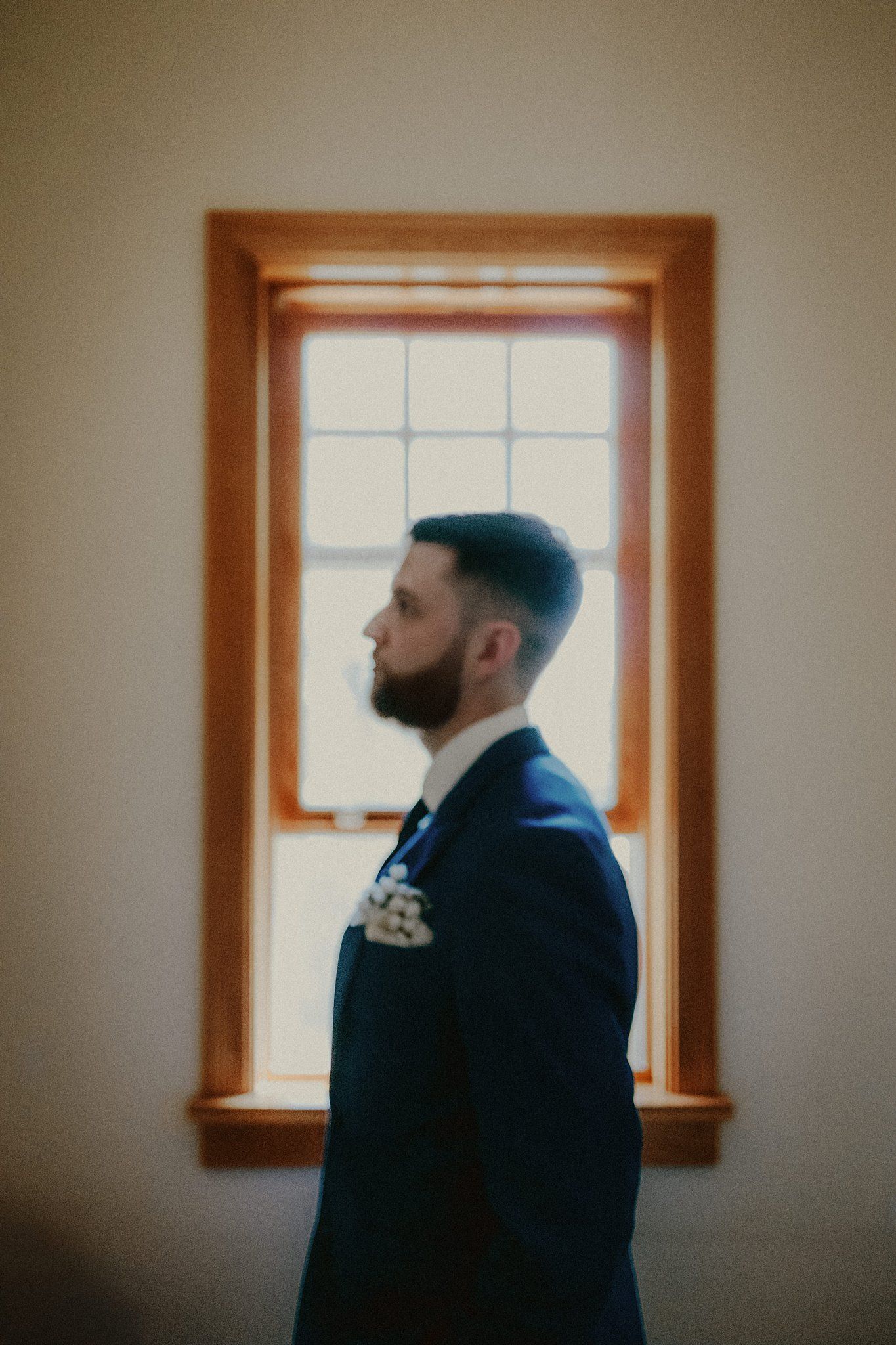 Silhouette of person in navy suit standing by window with wooden frame in moody lighting.