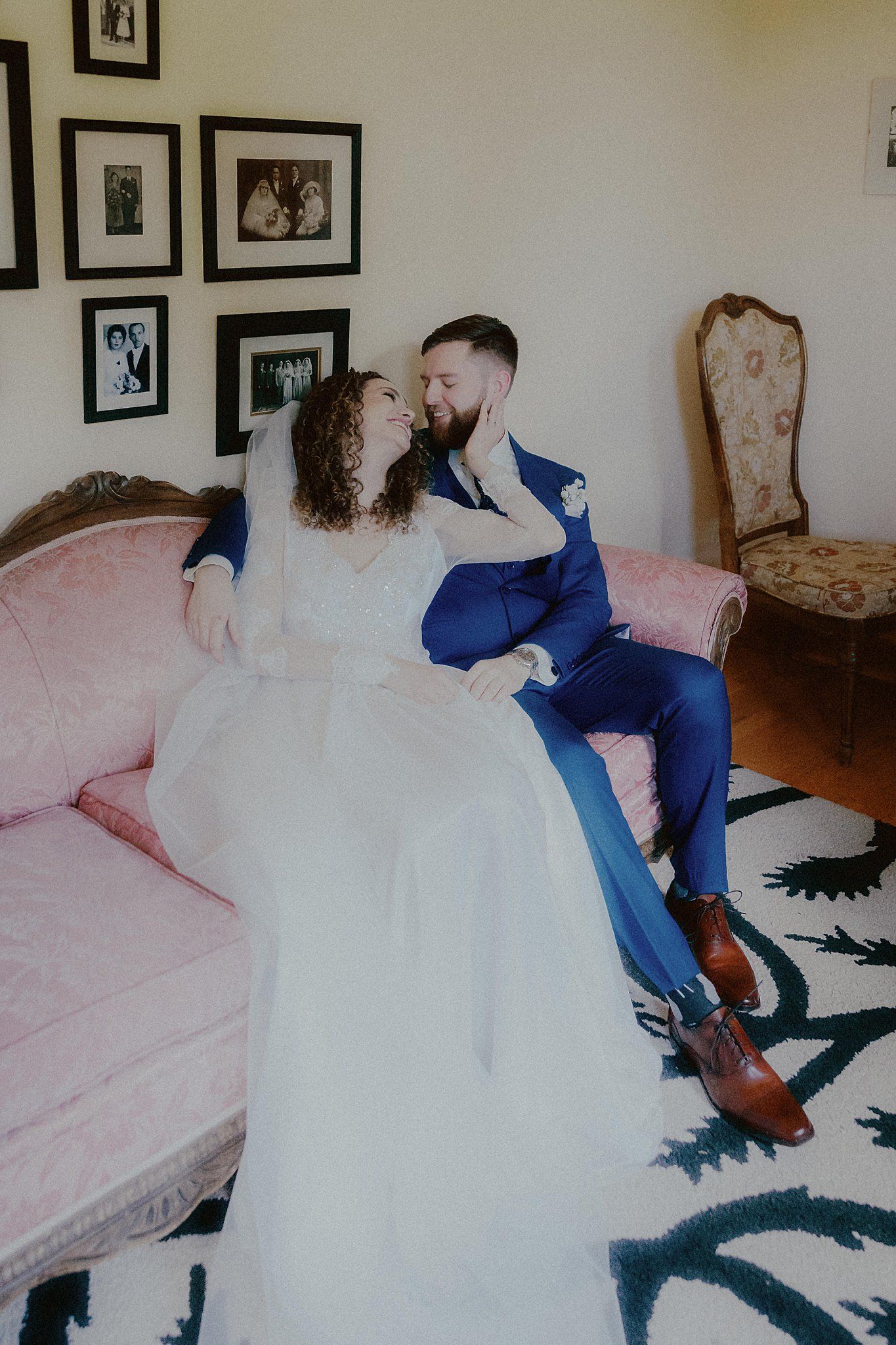 Couple posing on a vintage pink sofa with black and white photos on the wall.