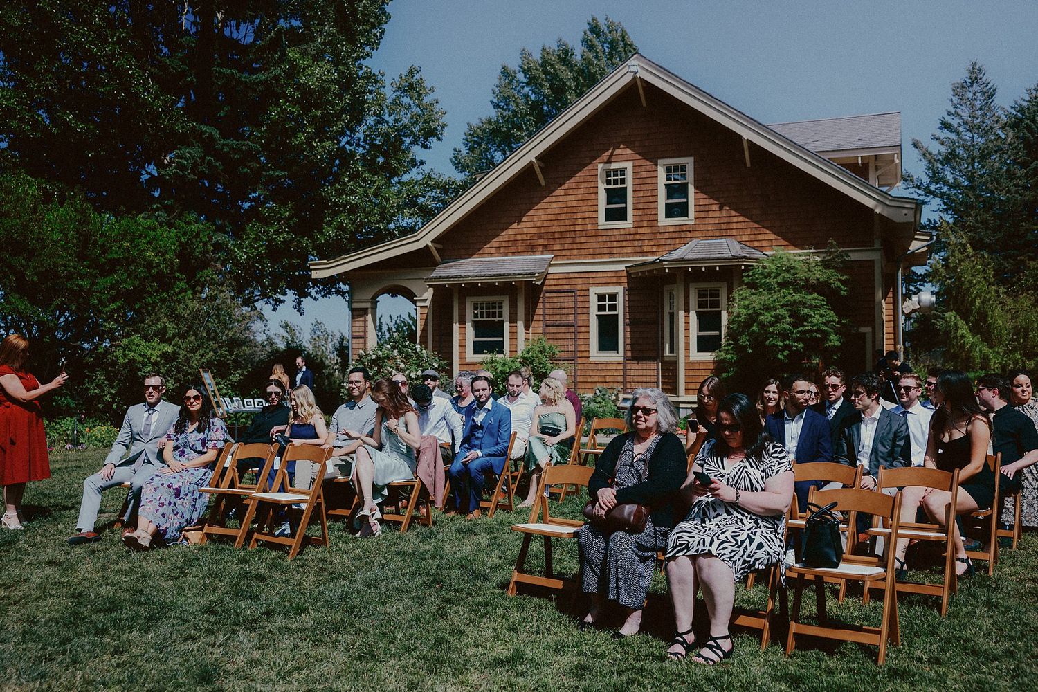 Wedding guests seated on wooden chairs outside rustic brown house for outdoor ceremony.