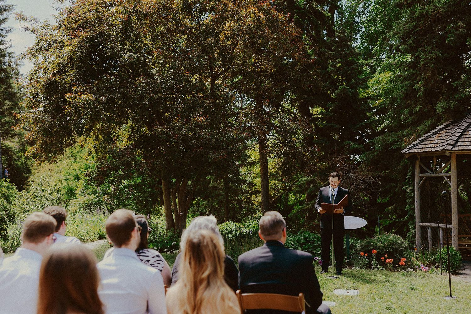 Outdoor wedding ceremony with guests seated facing gazebo among trees.