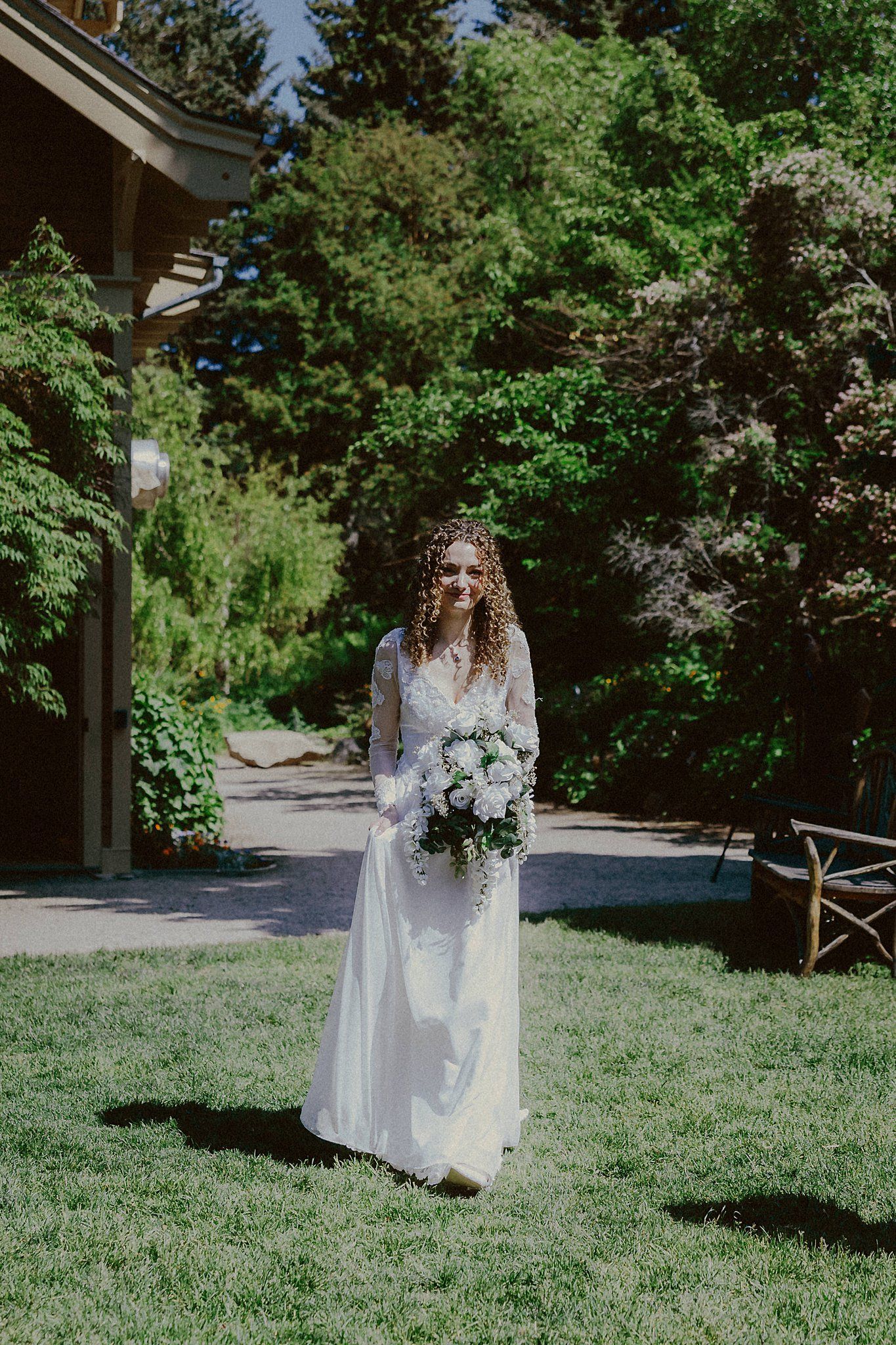 Bride holding white bouquet poses in garden setting wearing long sleeve lace gown.