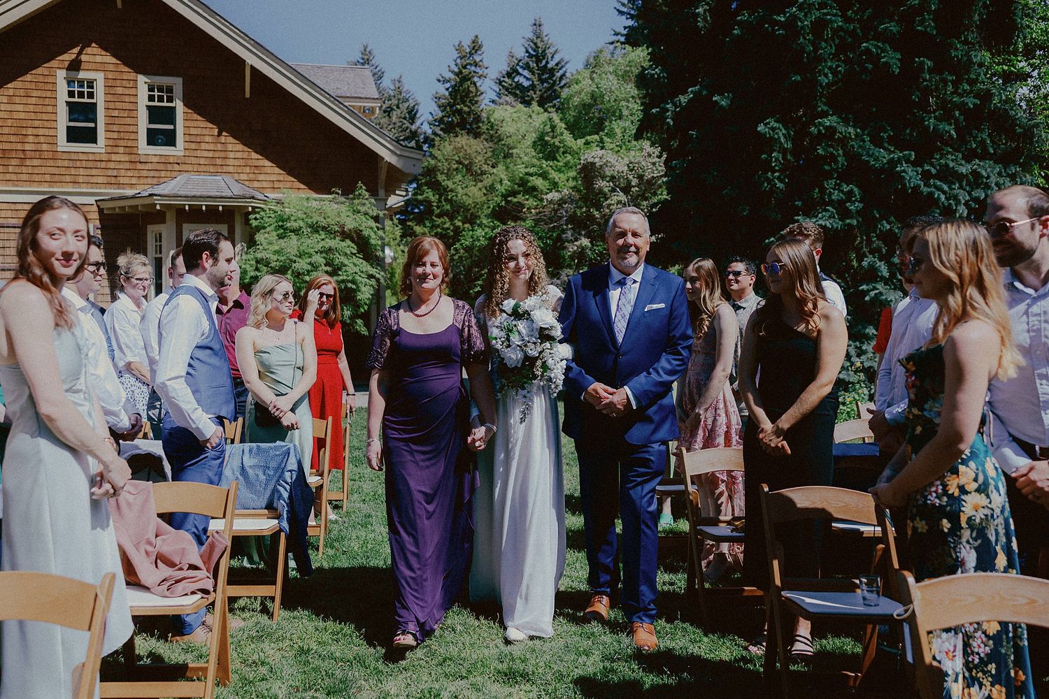 Wedding party and guests gather outside rustic wooden house for outdoor ceremony.