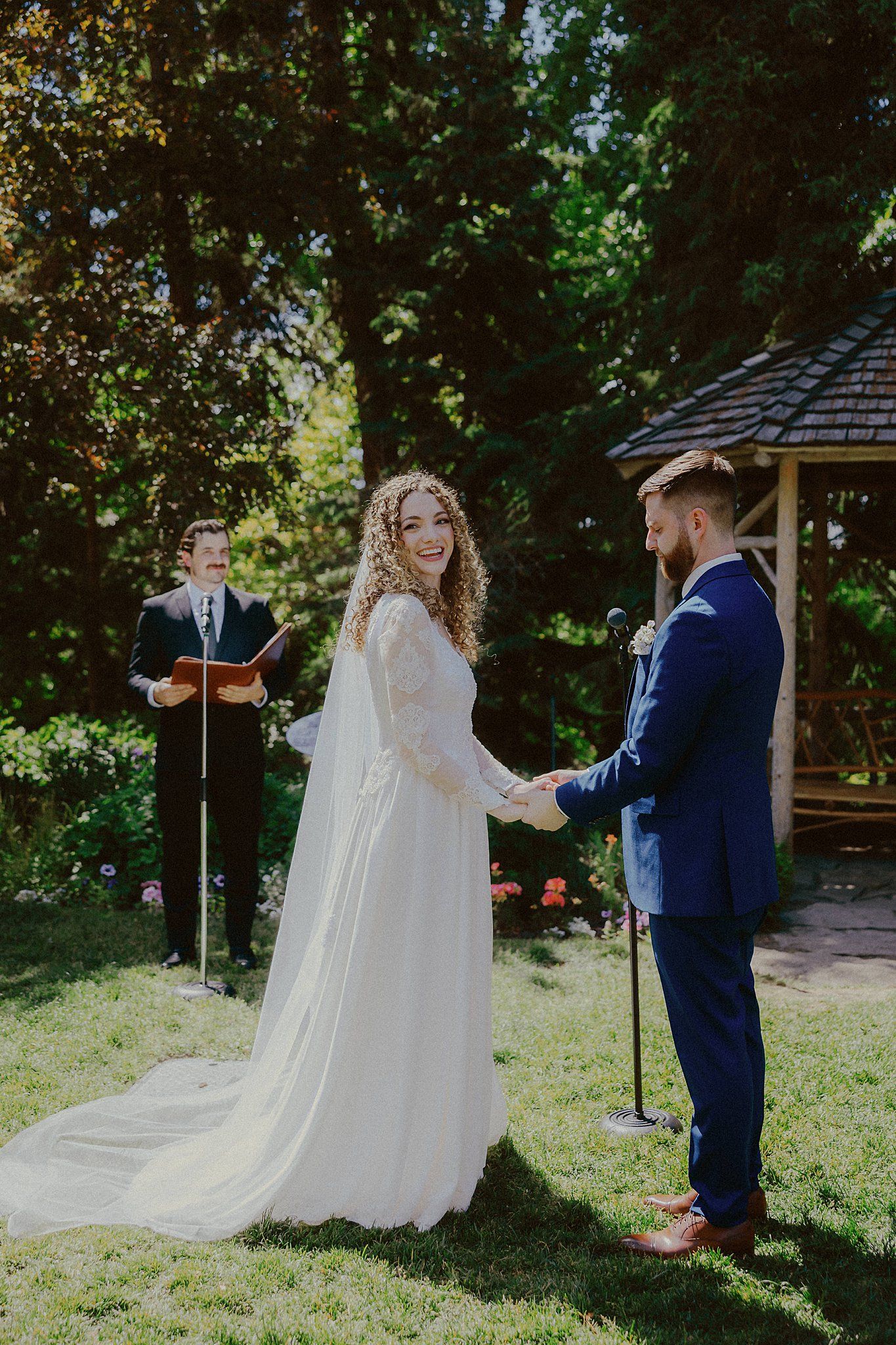 A wedding ceremony takes place in a lush garden setting with a white-dressed bride and navy-suited groom.