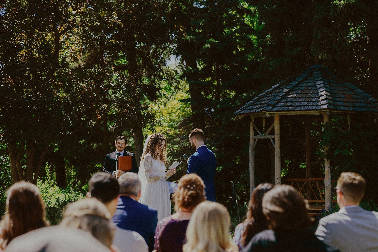 Wedding guests seated on wooden chairs watch the outdoor ceremony unfold.