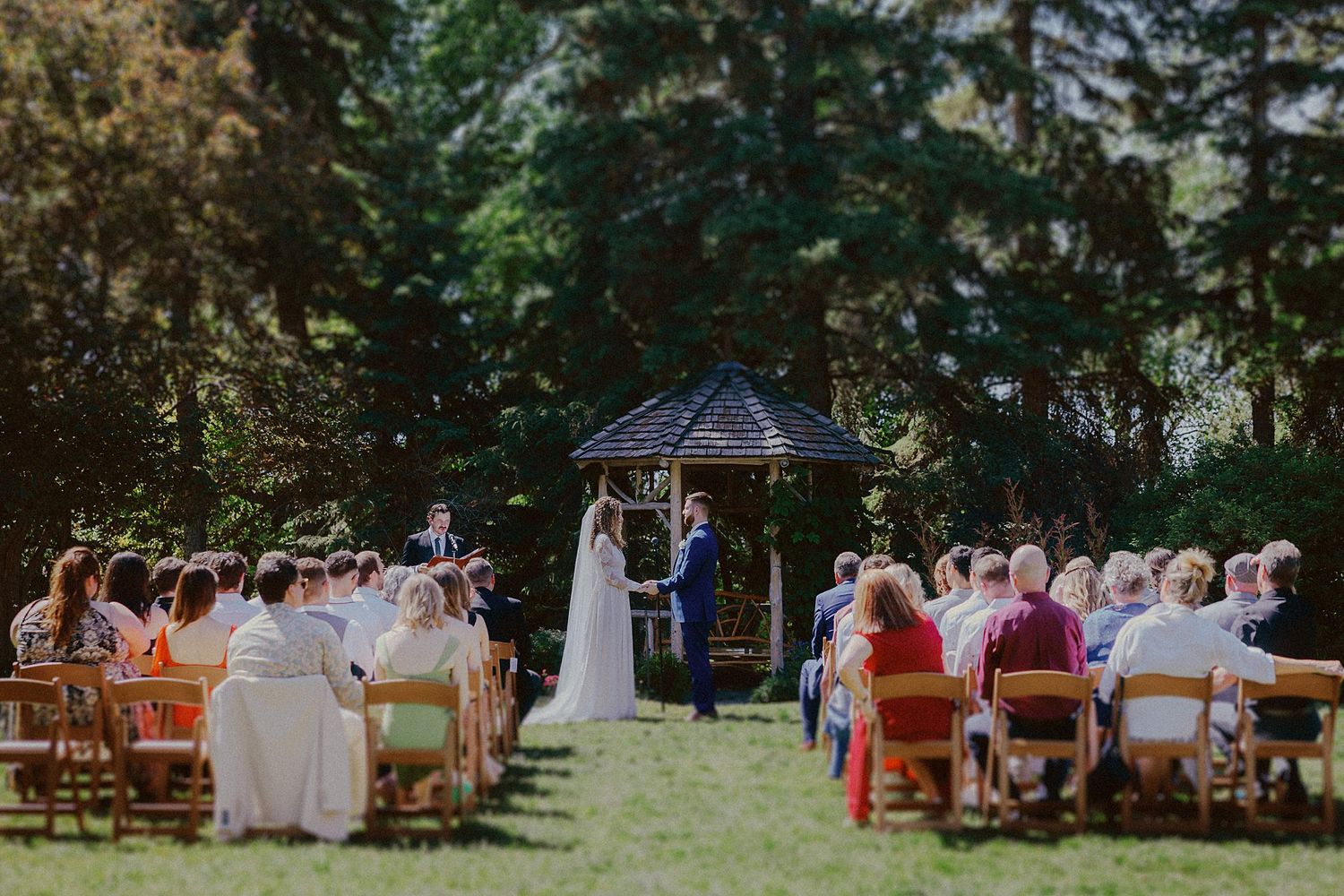 Panoramic view of an outdoor wedding ceremony with guests seated on wooden chairs.