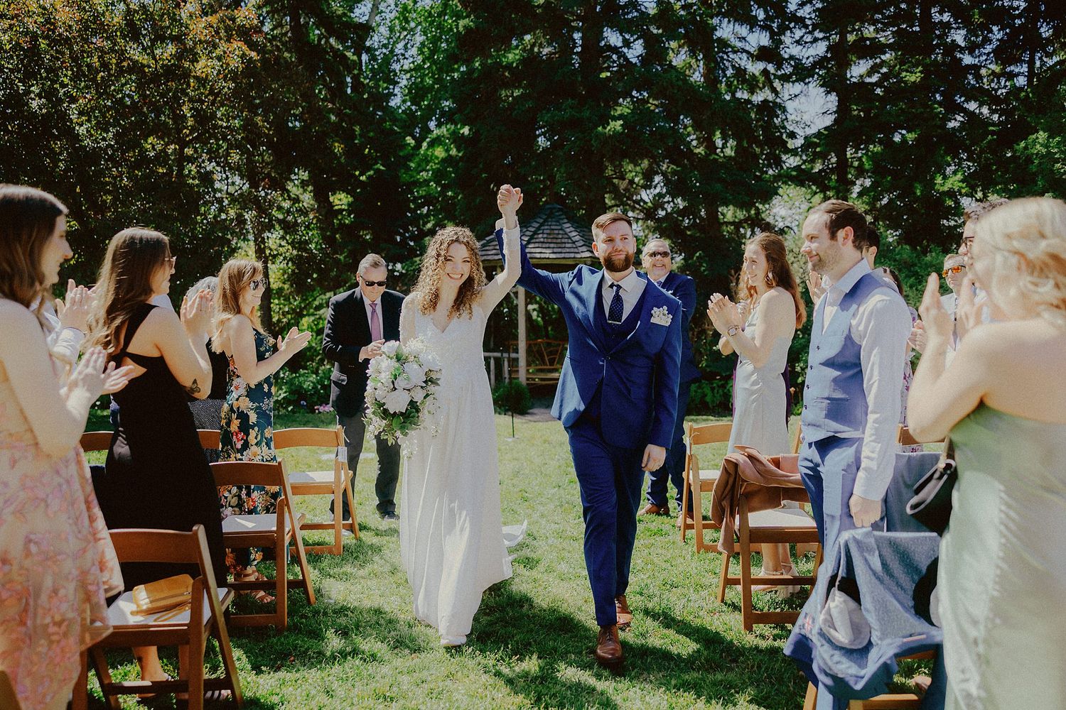 Wedding ceremony in progress with guests gathered under tall trees.