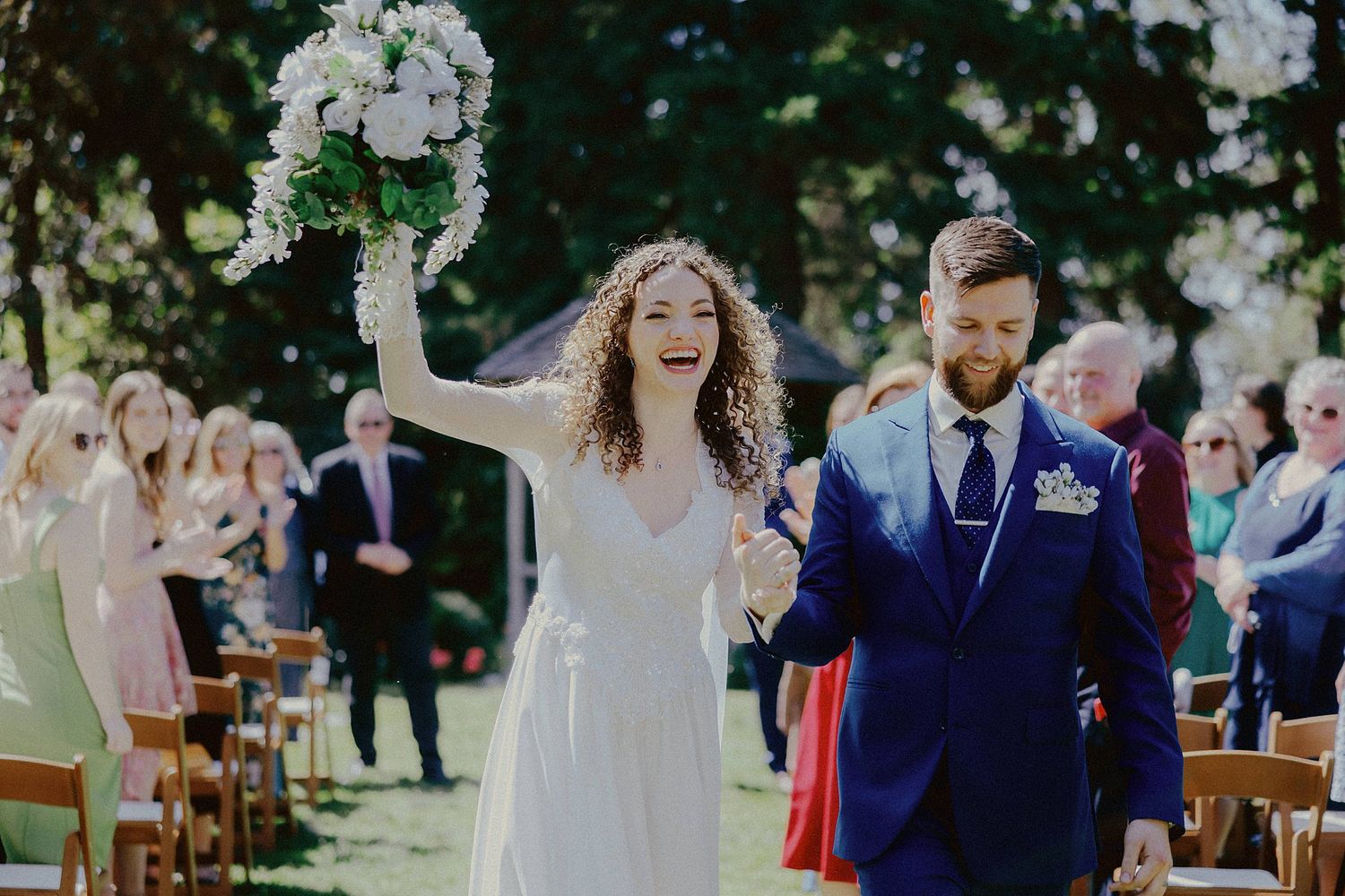 A joyful outdoor wedding ceremony with guests seated on wooden chairs surrounded by lush greenery and floral arrangements.