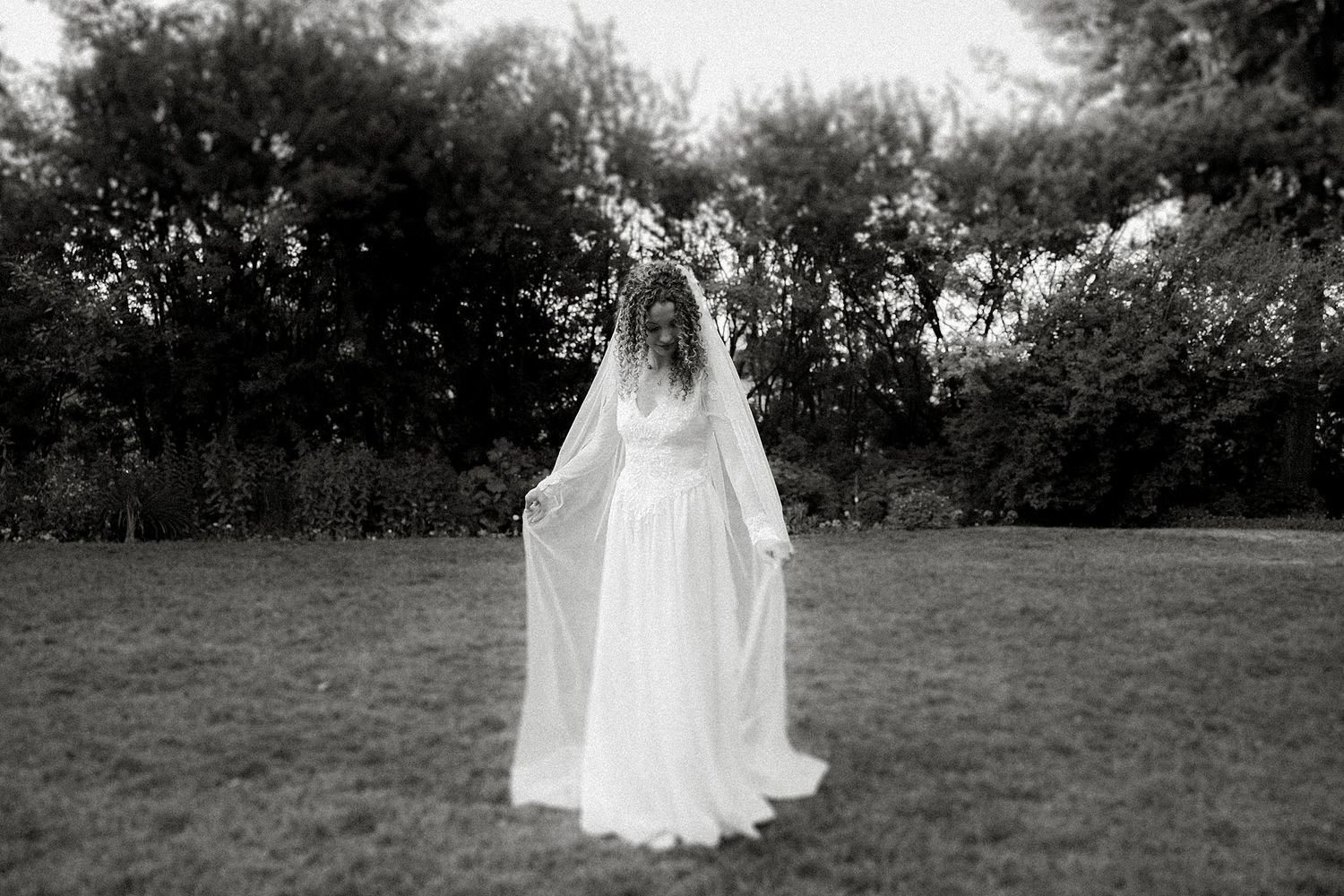 A dreamy black and white portrait of a bride in a flowing dress and veil standing in a garden.
