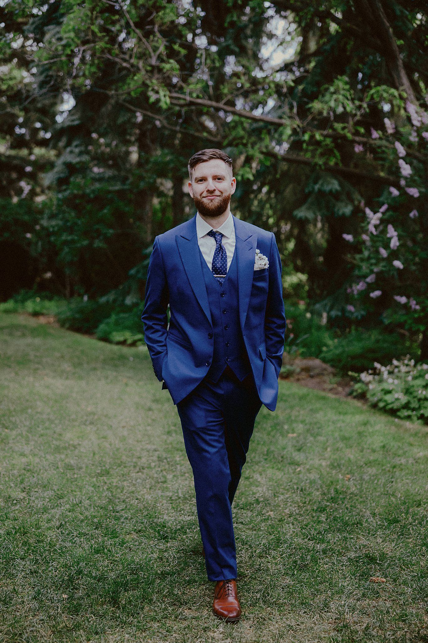 Wedding party members in navy blue suits walking on grass with greenery in background.
