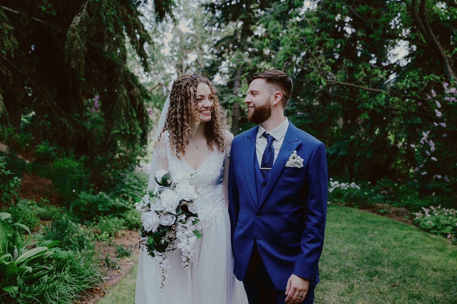 A couple in navy blue wedding attire embraces while standing in a lush green garden setting.